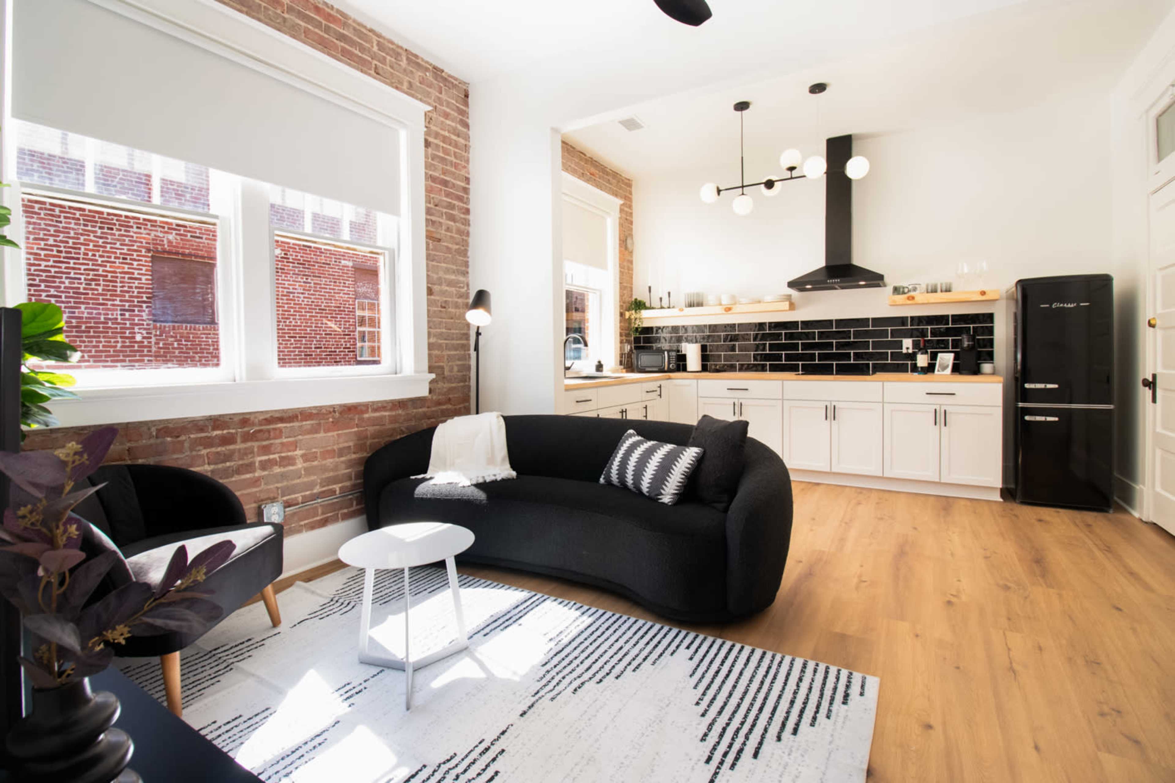 A modern living space featuring a black curved sofa, a round white side table, and a kitchen area with white cabinets and a black backsplash.