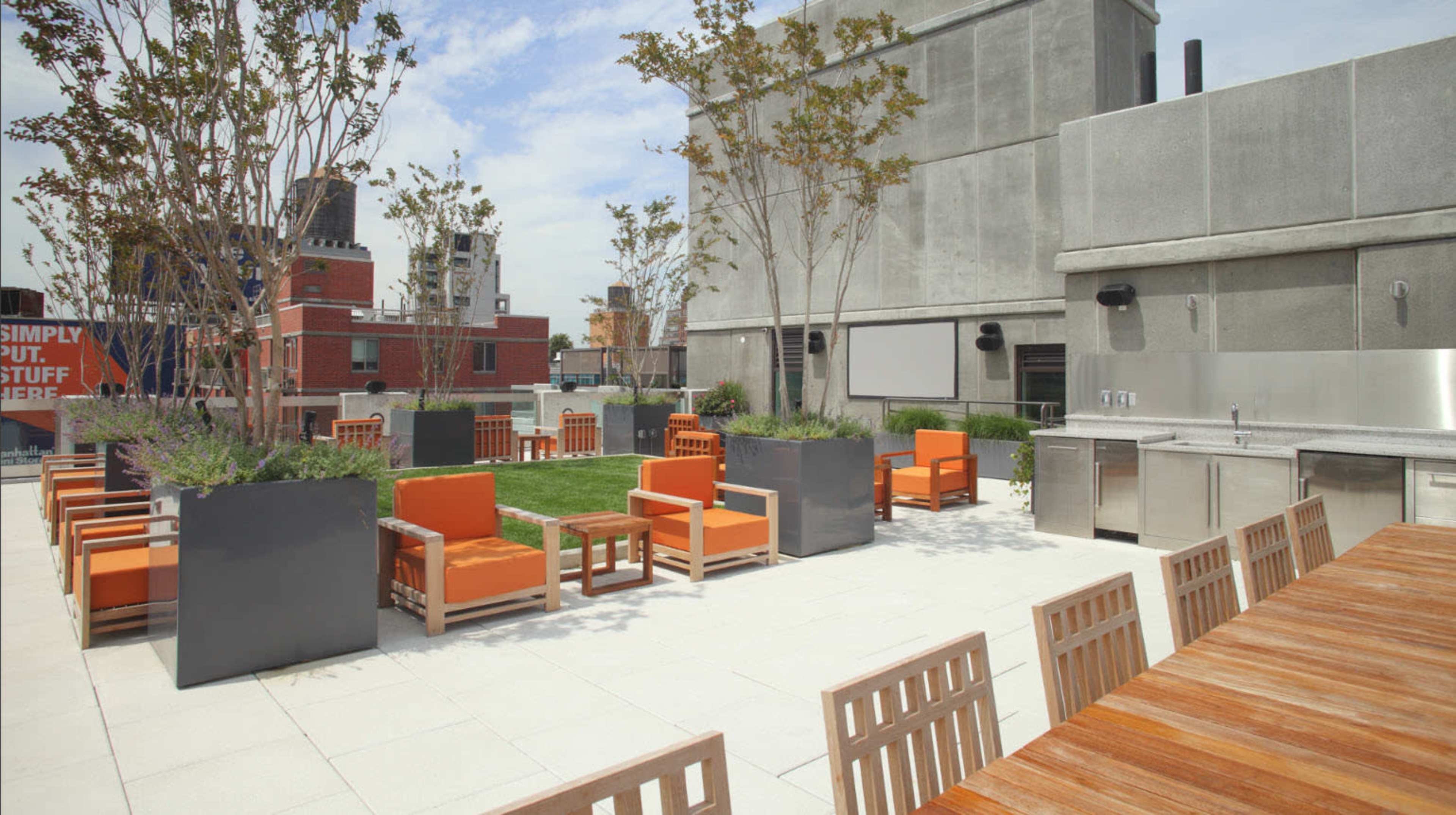 A rooftop terrace featuring orange chairs, wooden tables, and planters with greenery.