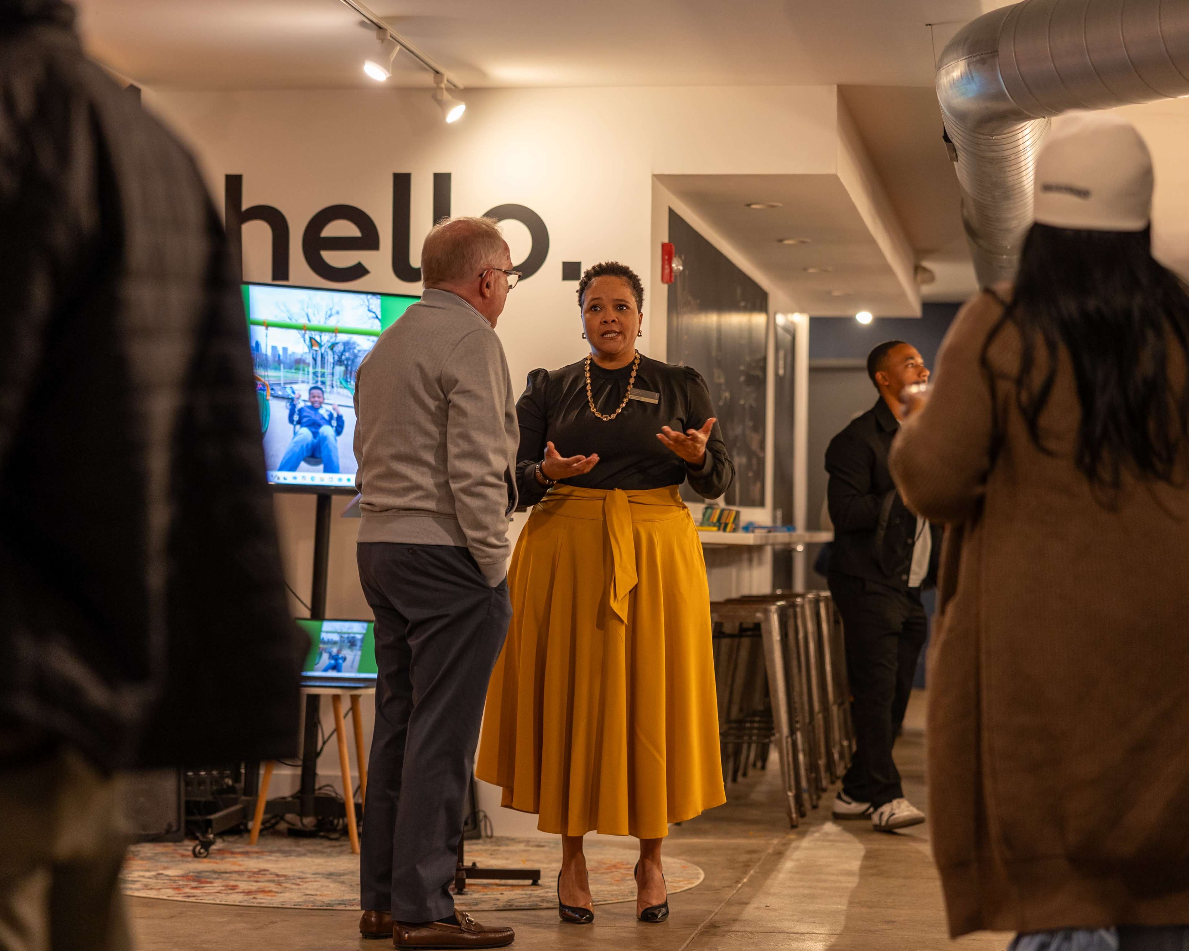 A woman in a black top and yellow skirt engages in conversation with a man in a gray sweater, while others socialize in a modern indoor space.