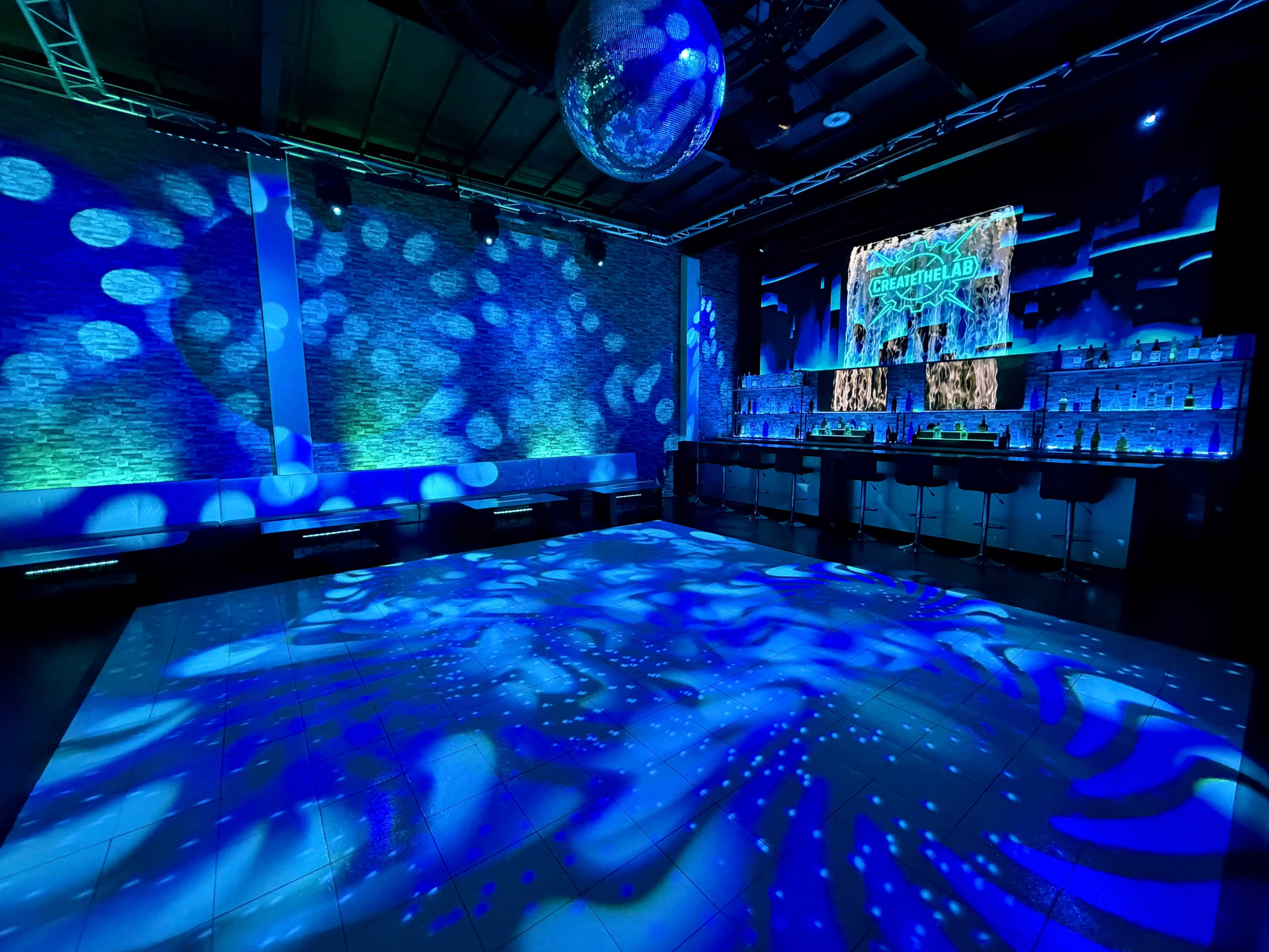 The image shows a dimly lit bar area with a shimmering disco ball, illuminated by blue lights and patterns projected on the floor and walls.