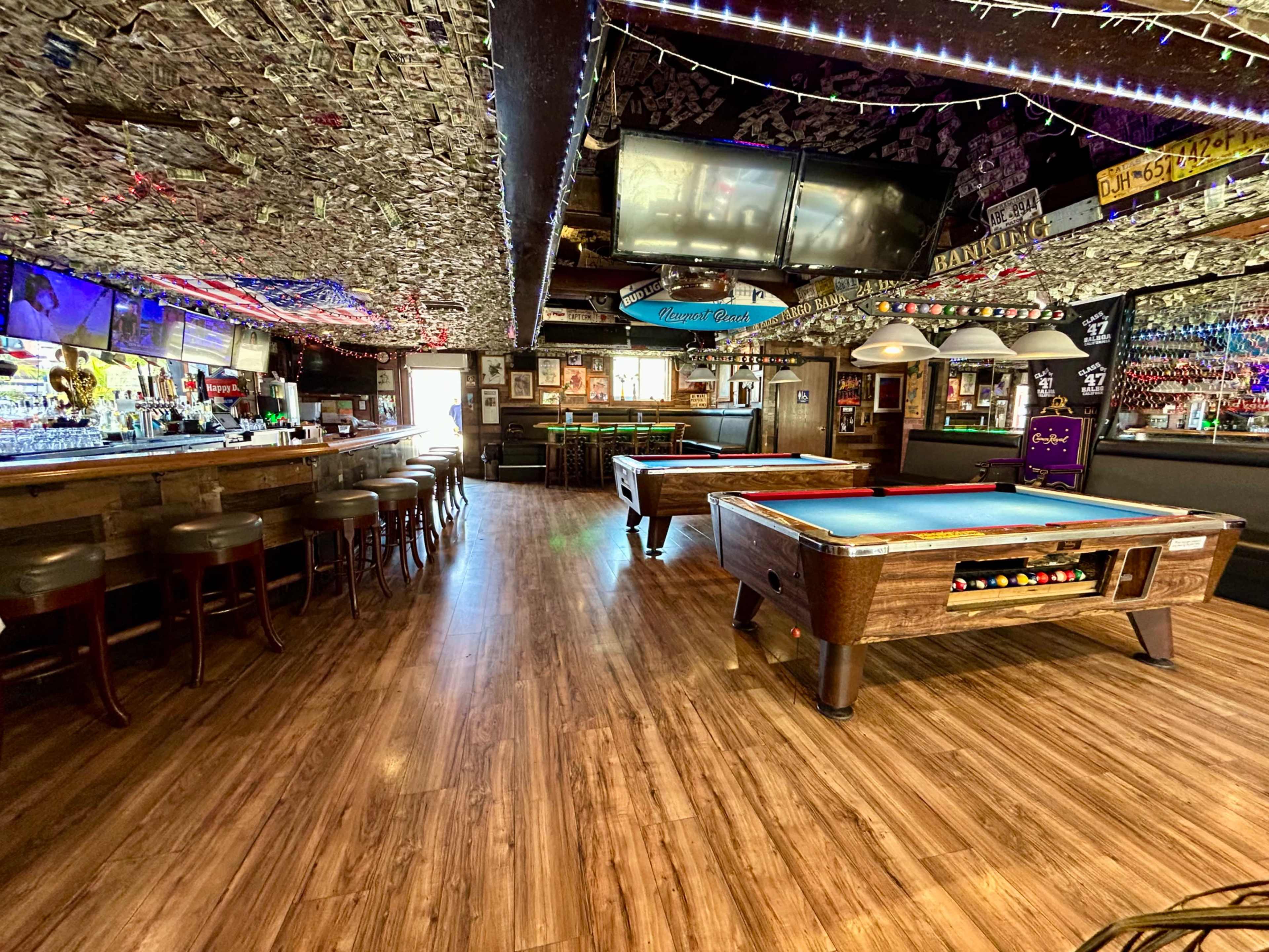 The image shows the interior of a rustic bar with two pool tables and a long bar counter, adorned with decorations and dollar bills on the ceiling.