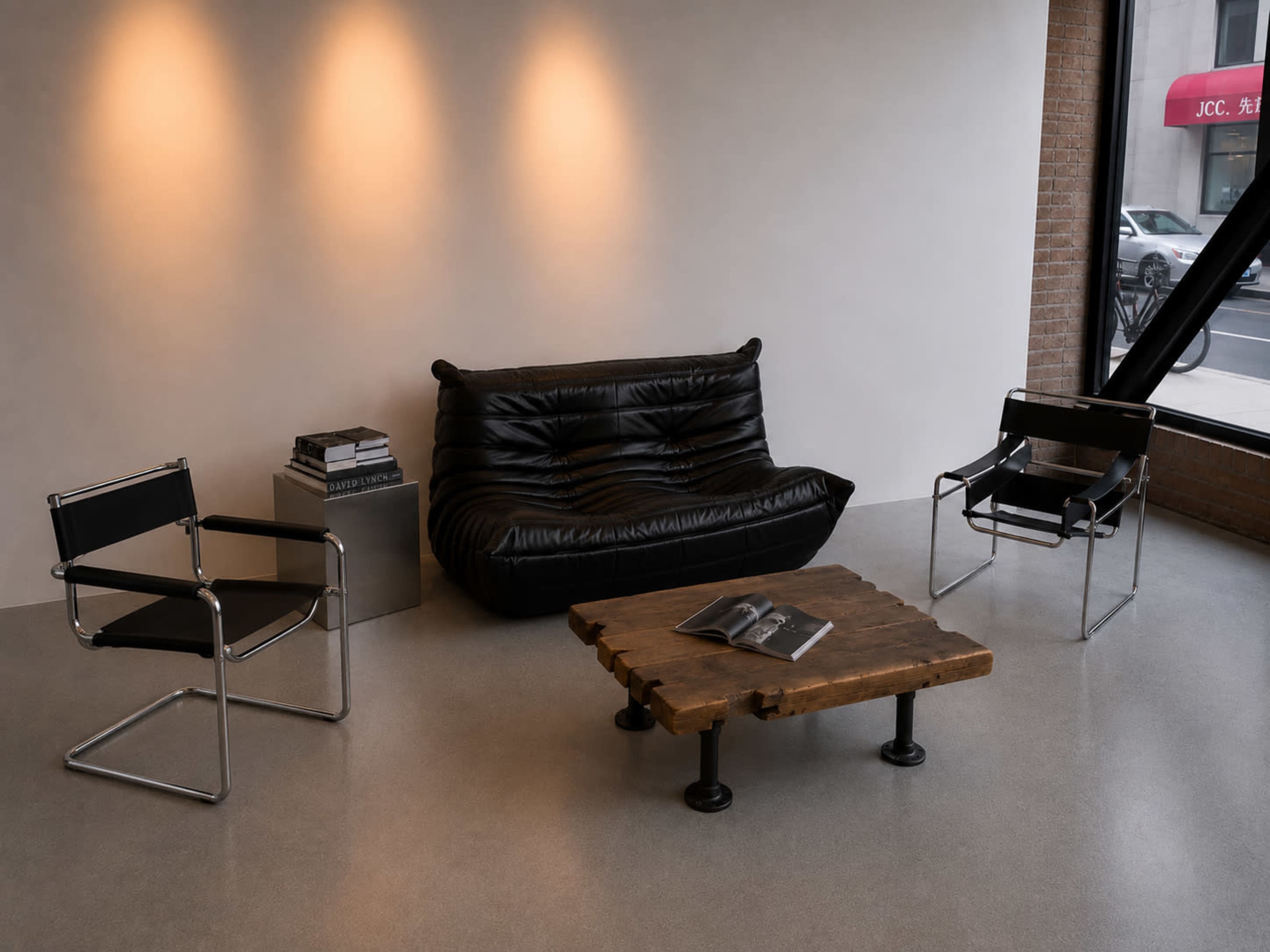 The image features a minimalist interior with a black sofa, two black chairs, a wooden coffee table, and a stack of books.