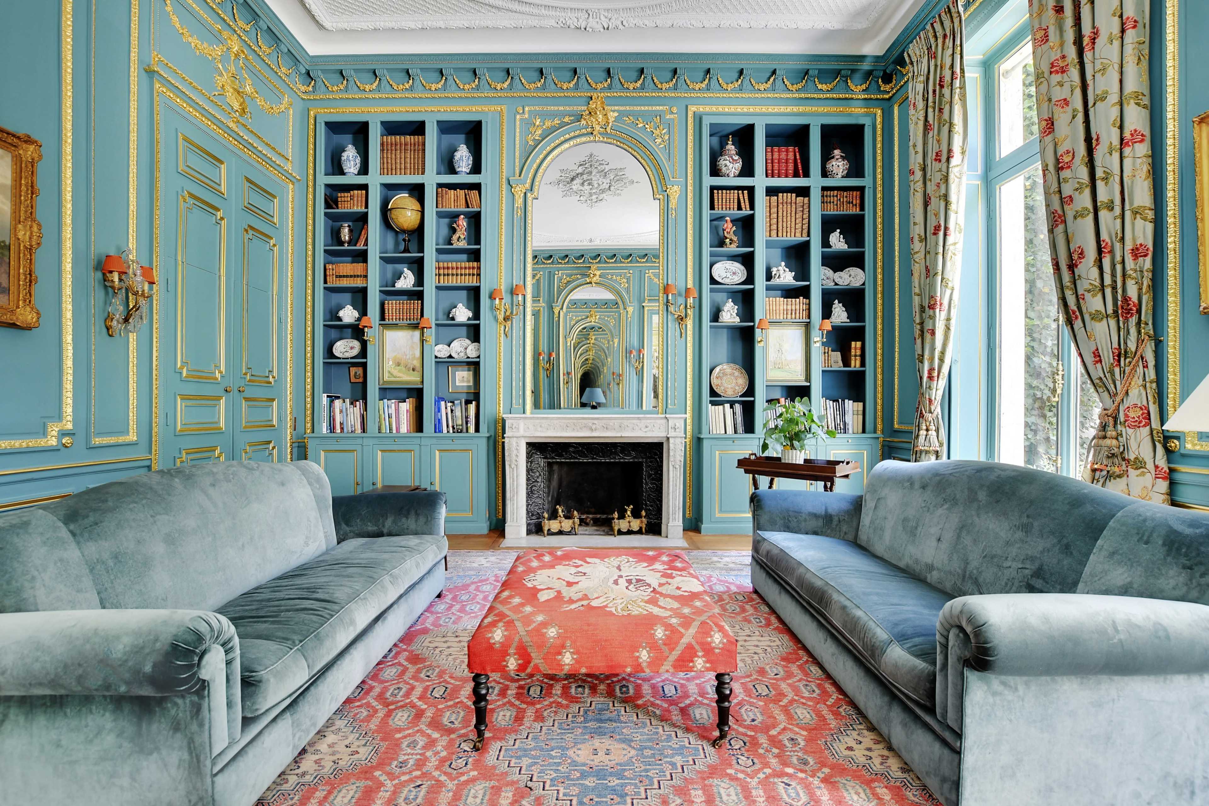The room features two velvet sofas facing a central ornate rug, with a large bookshelf and a decorative fireplace against the blue walls.