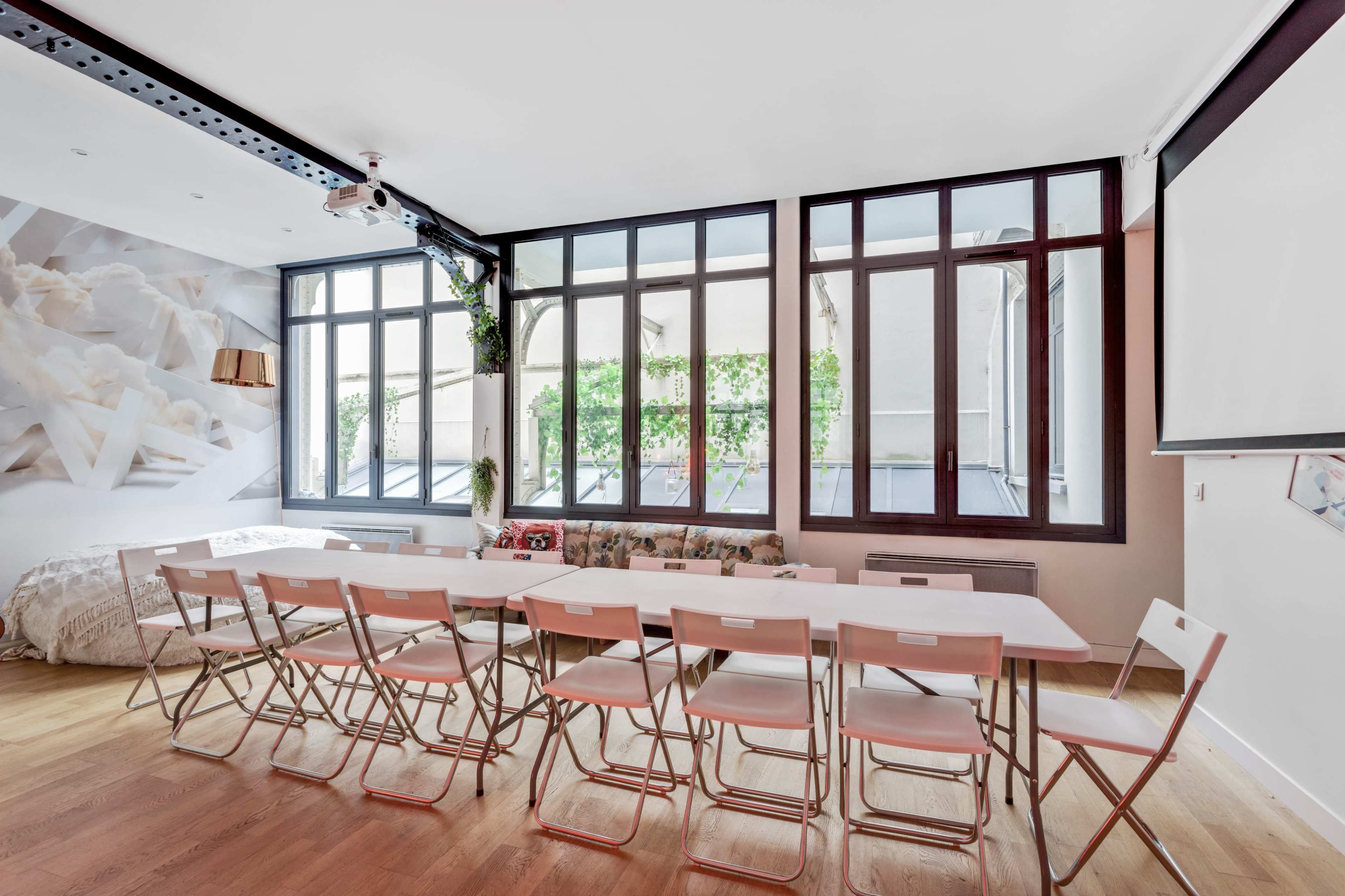 The image shows a spacious room with large windows, a long table with several folding chairs arranged neatly, and a cozy seating area in the background.