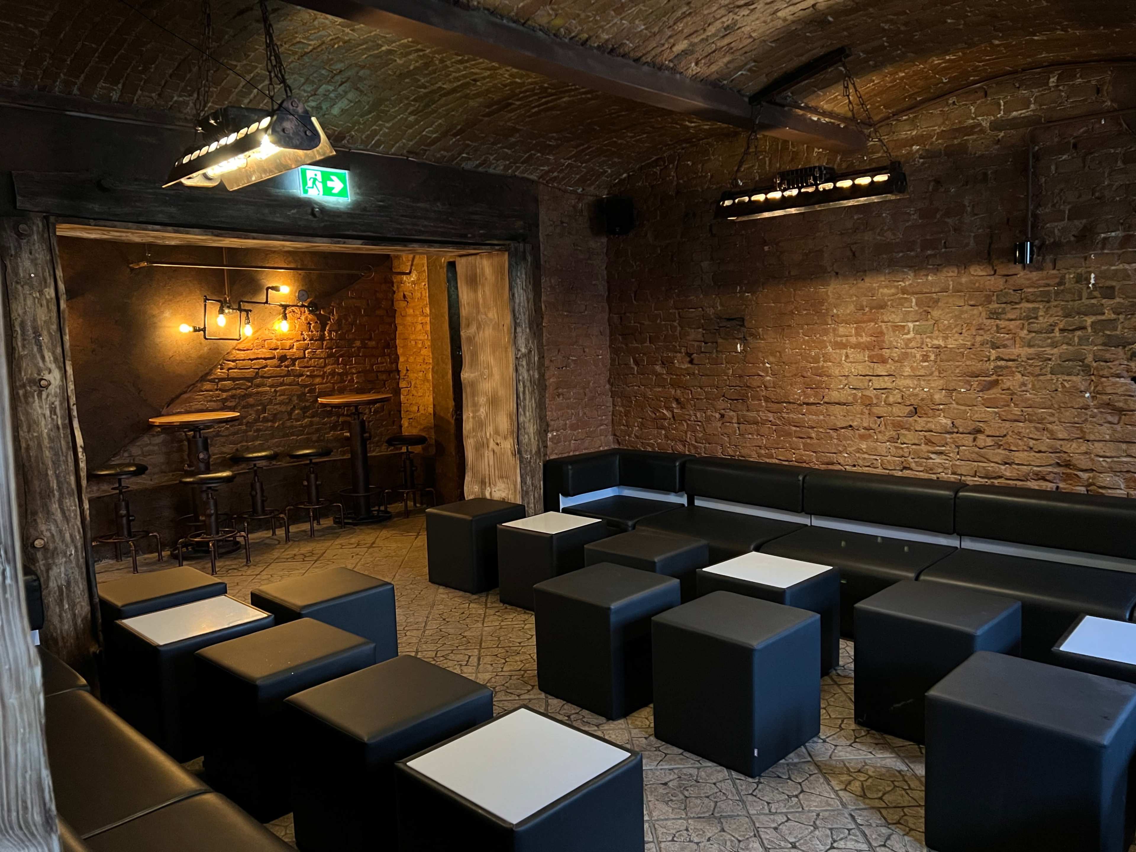 A dimly lit room with brick walls, featuring black seating cubes arranged in a social layout, and a small bar area visible in the background.