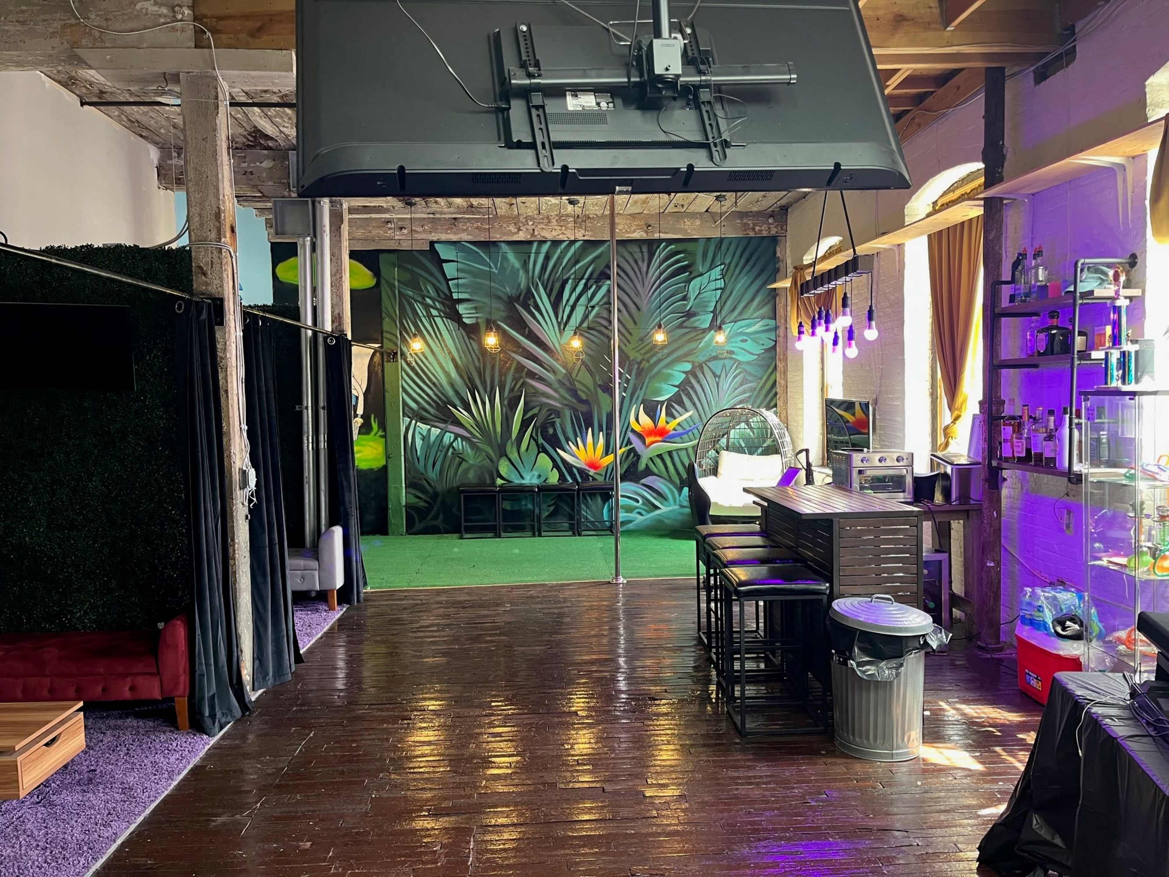 The interior of a spacious venue features a large wall mural of tropical plants, a bar area with seating, and subtle lighting.