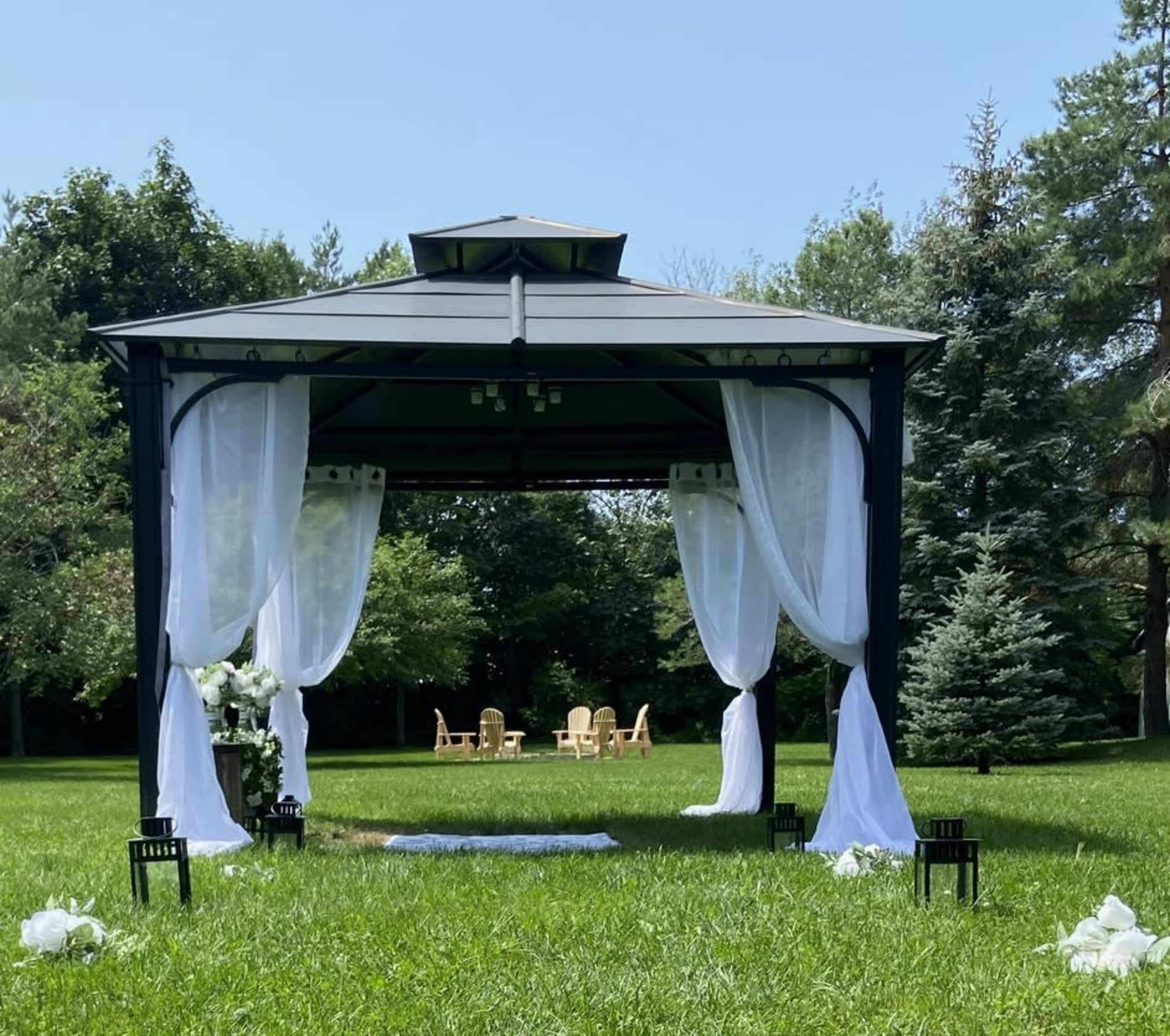A black gazebo with white drapes is set up on a grassy area, surrounded by decorative chairs and flower arrangements for an event.