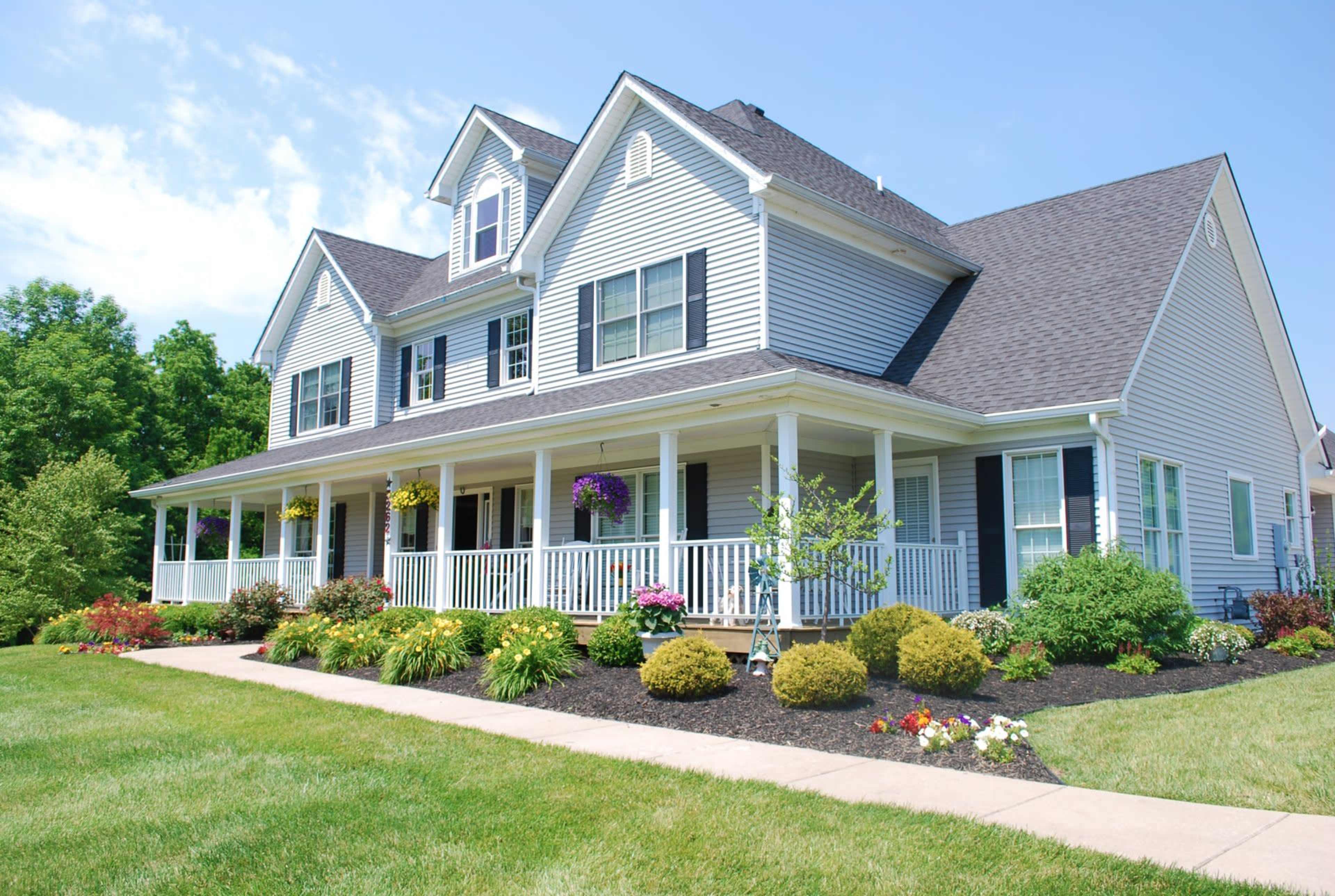 A large, two-story suburban house with a covered front porch and landscaped garden featuring colorful flowers and shrubs.