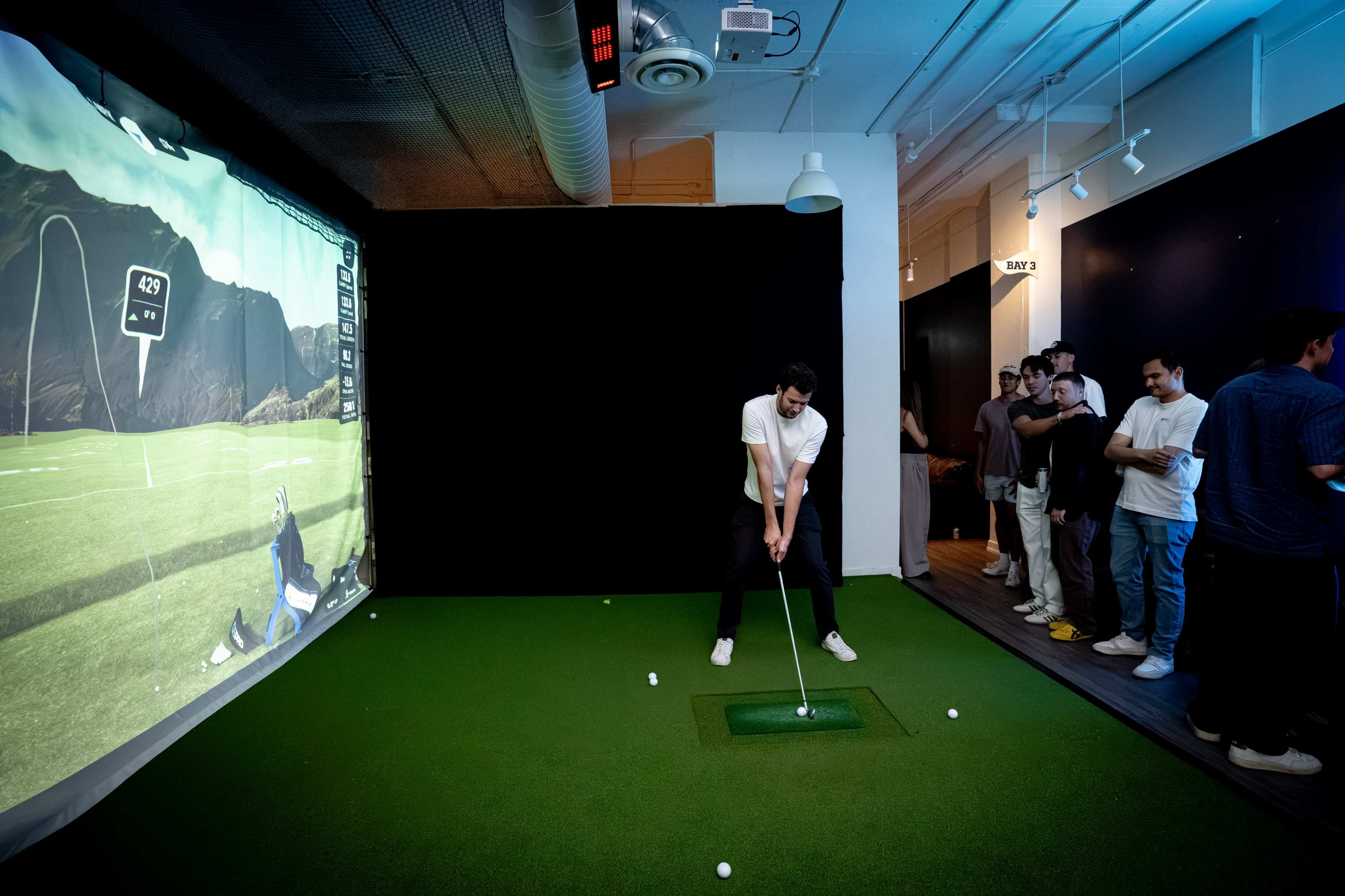 A group of people watches as one individual prepares to take a golf shot in a simulated indoor driving range.