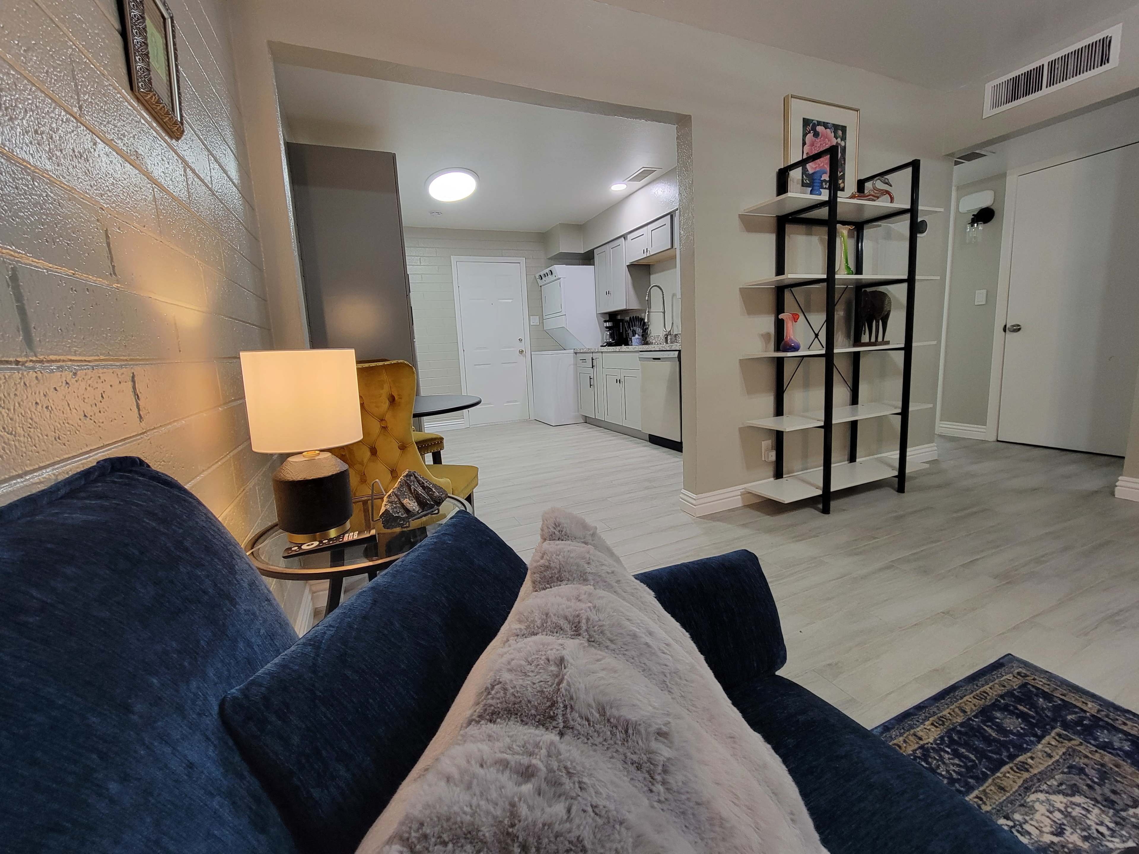 The image shows a cozy living room area with a blue couch, a lamp, and a bookshelf, leading into an open kitchen space.