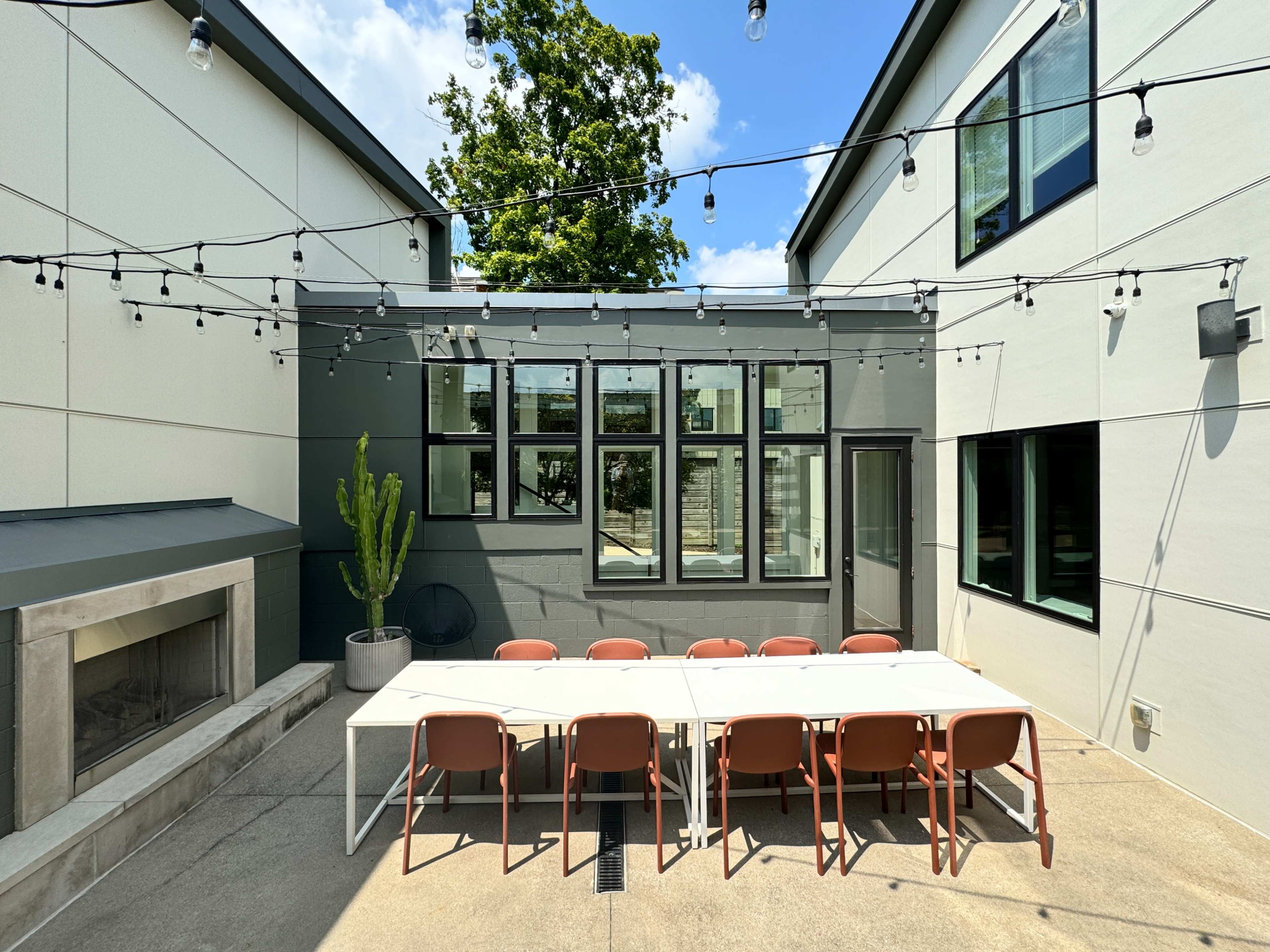 The image shows a modern outdoor patio area with a long white table, eight orange chairs, and string lights overhead, surrounded by buildings with large windows.