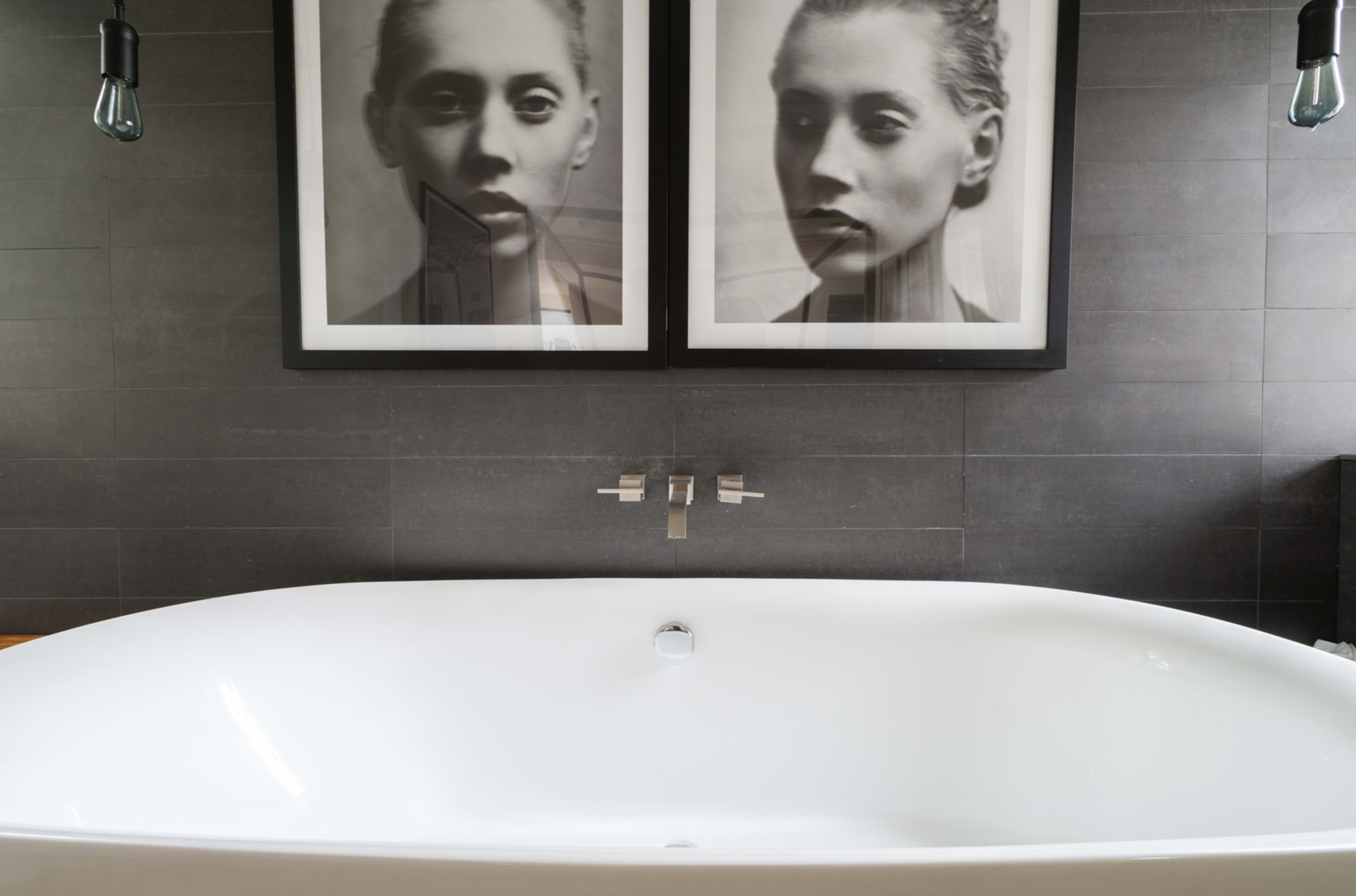 The image shows a modern bathroom featuring a freestanding white bathtub and two large black-and-white portraits mounted on the wall above it.