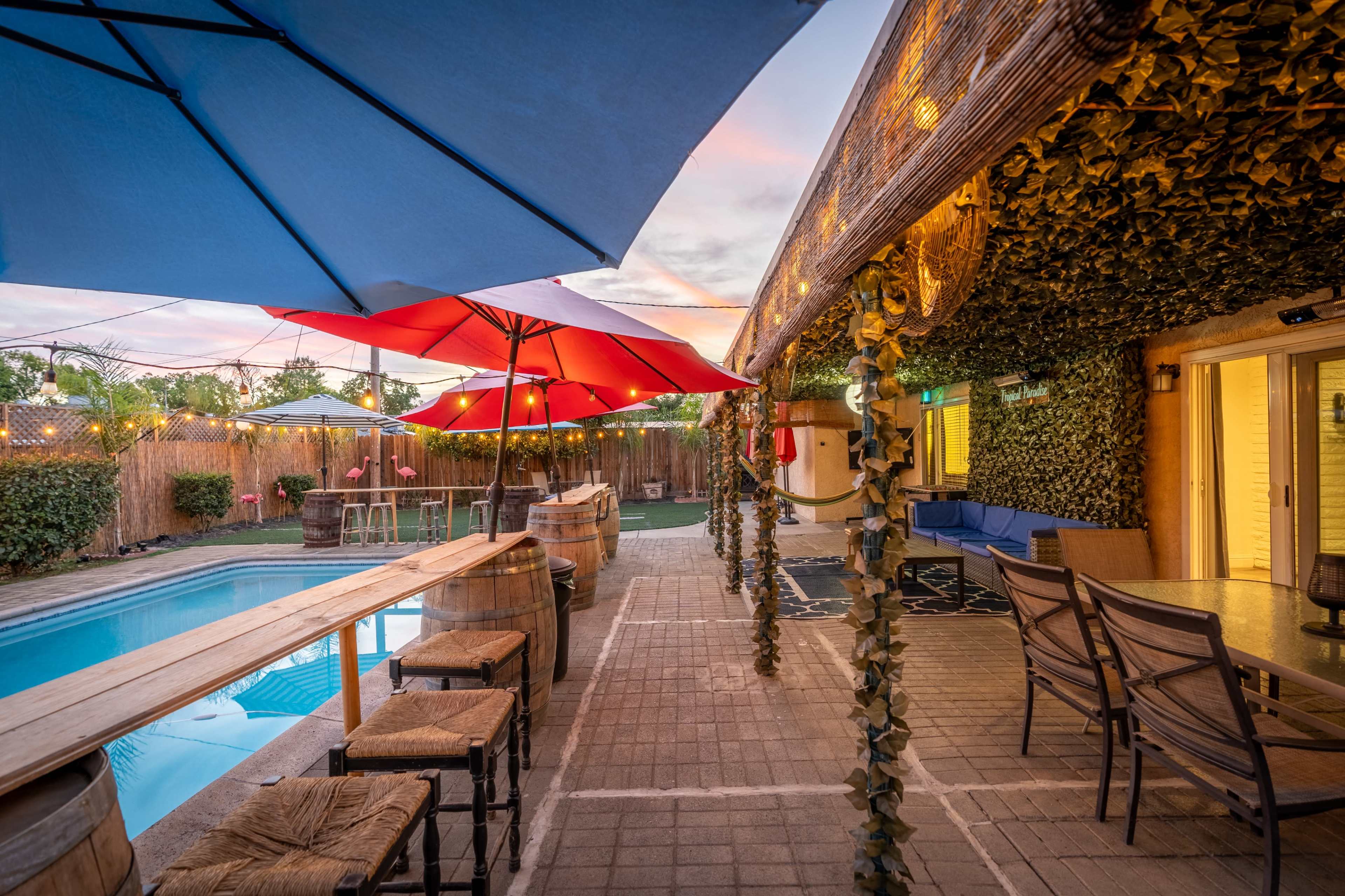 A swimming pool area with a bar, blue and red umbrellas, and seating surrounded by greenery and string lights.
