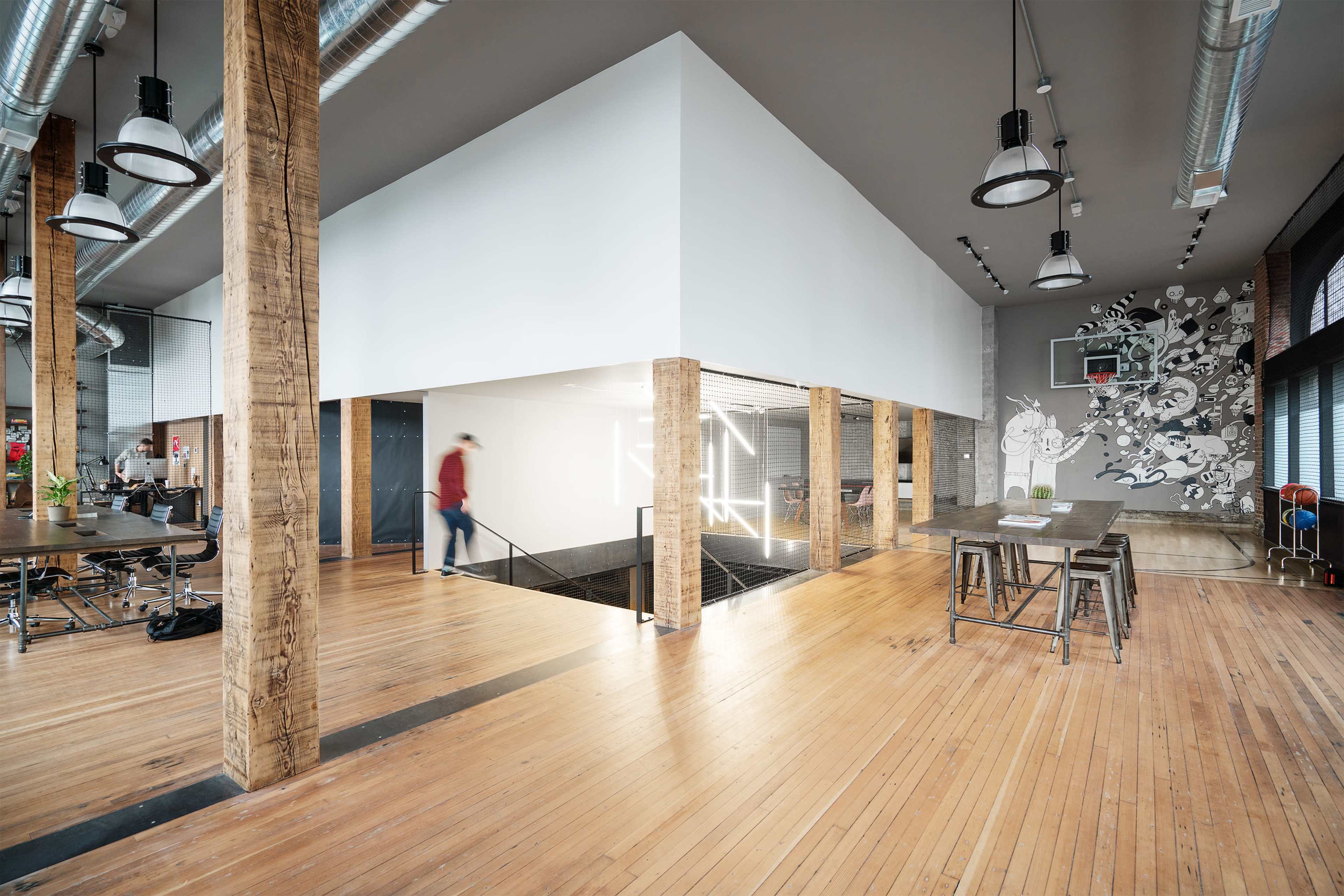 The image shows an open office space with wooden columns, a white-cube structure in the center, and a mural on the wall, featuring tables and a basketball hoop in the background.