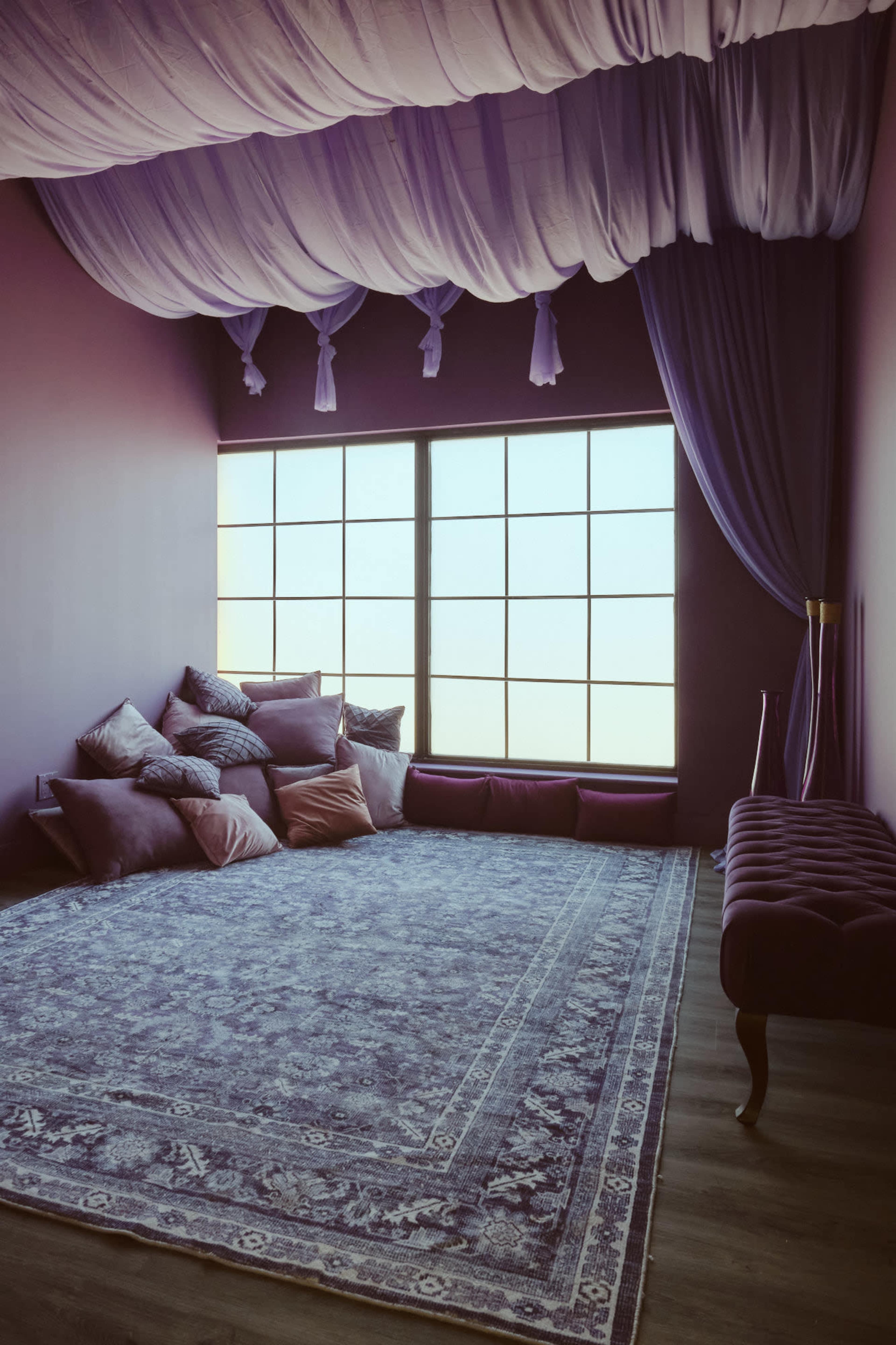 The image depicts a spacious, softly lit room with a large window, adorned with plush pillows and a decorative rug on the wooden floor.