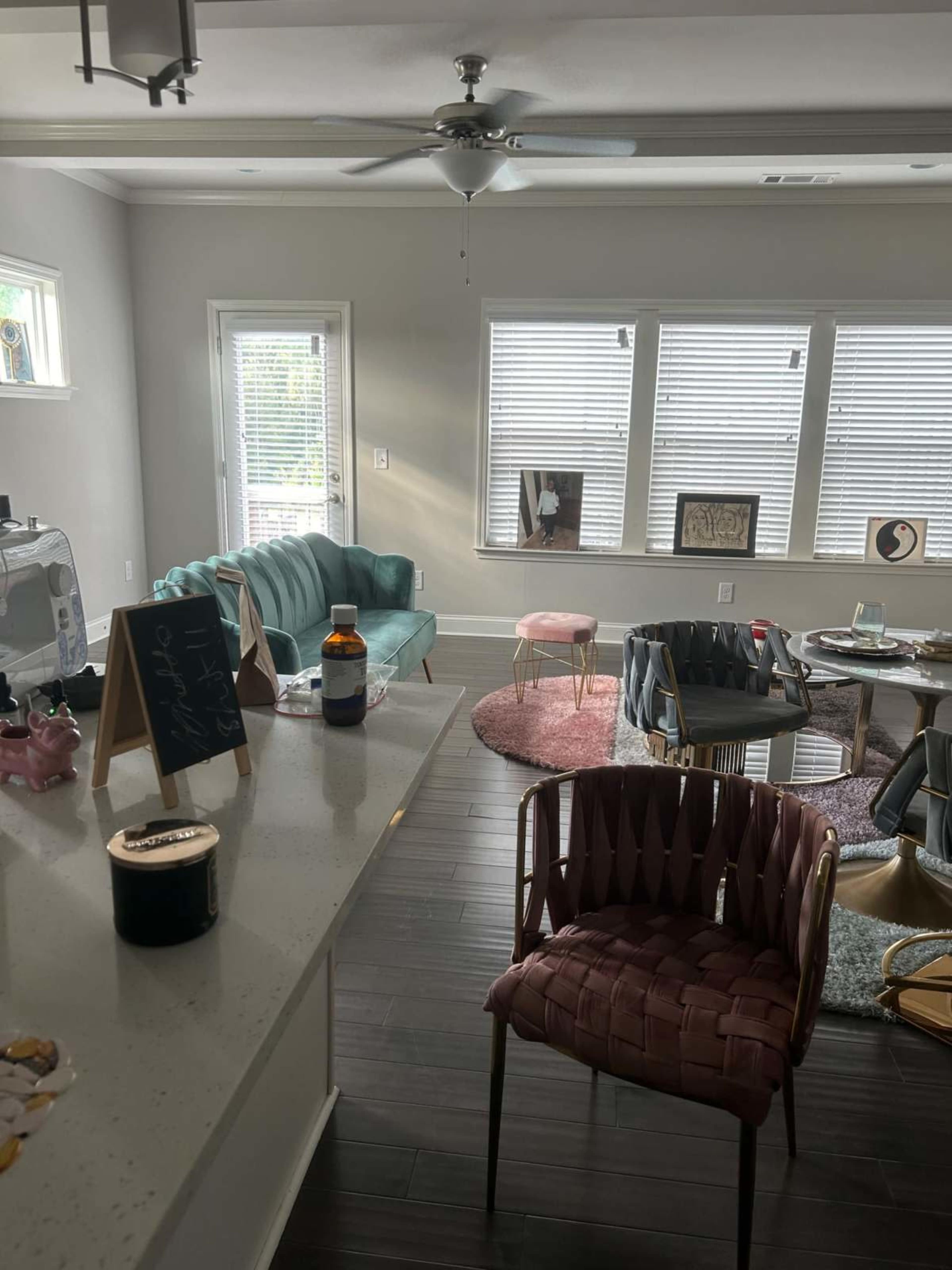 The image shows a modern living room with a teal sofa, a pink stool, and several decorative chairs arranged around a low coffee table, illuminated by natural light from windows.