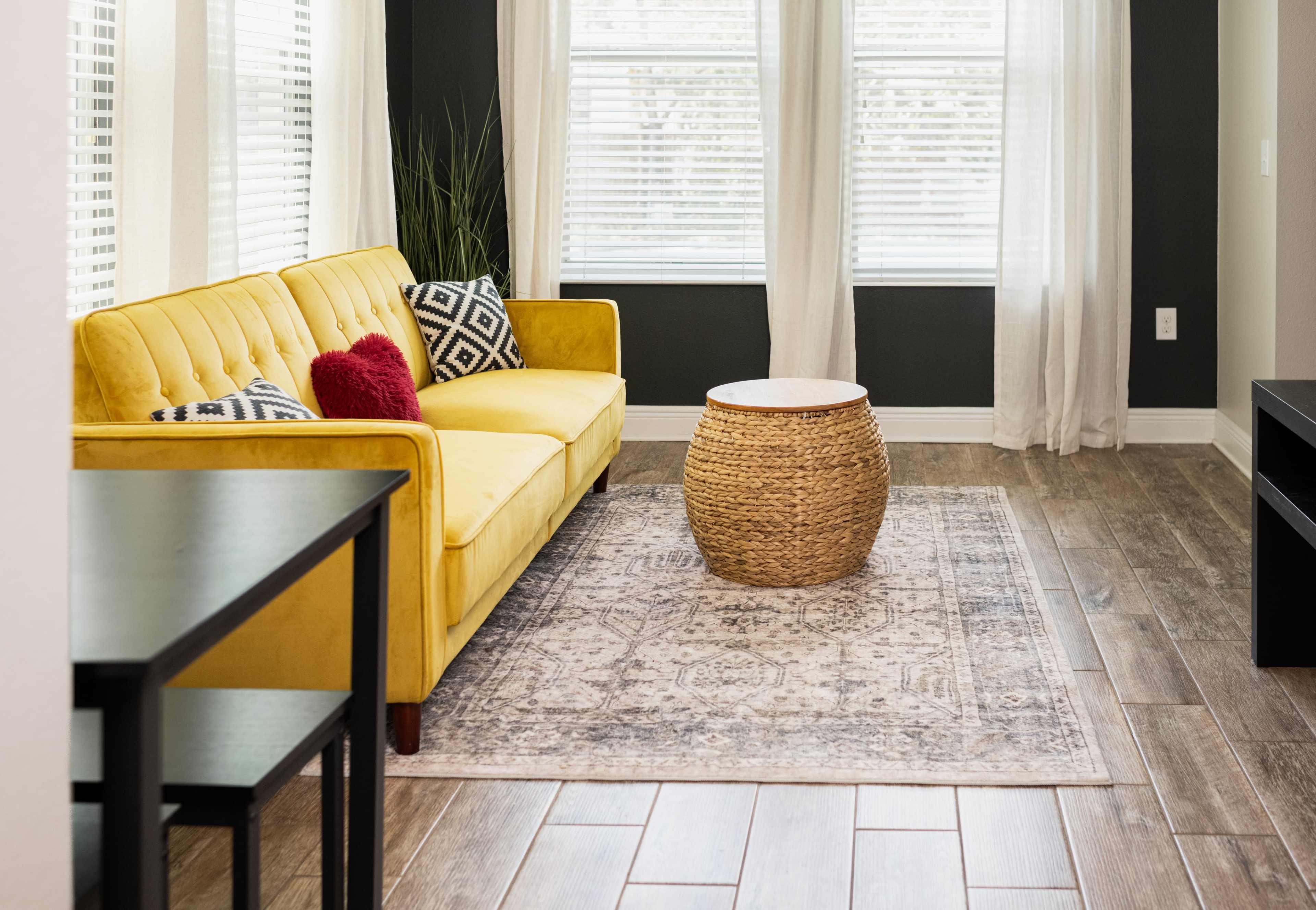 A bright yellow sofa is positioned in a room with large windows, a round woven coffee table, and a patterned area rug.
