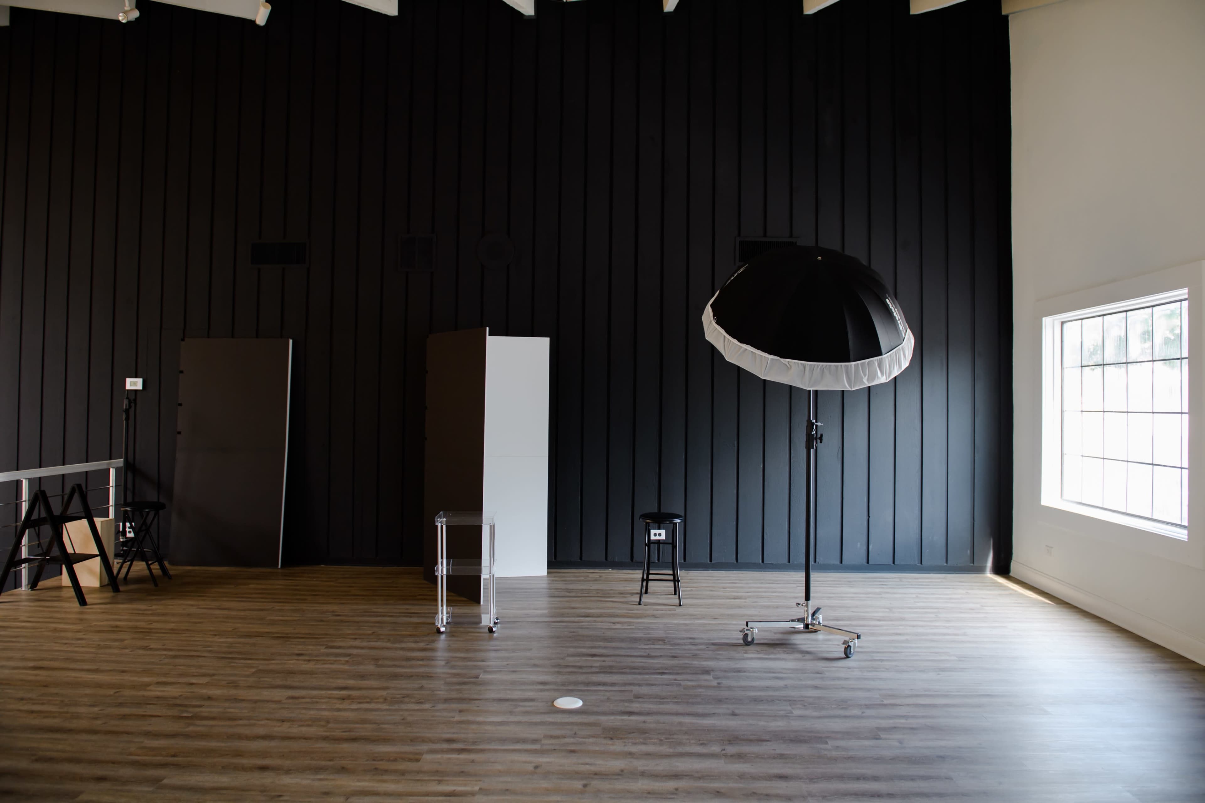 An empty photography studio with a black wooden wall, a large umbrella softbox on a stand, and minimal furniture.