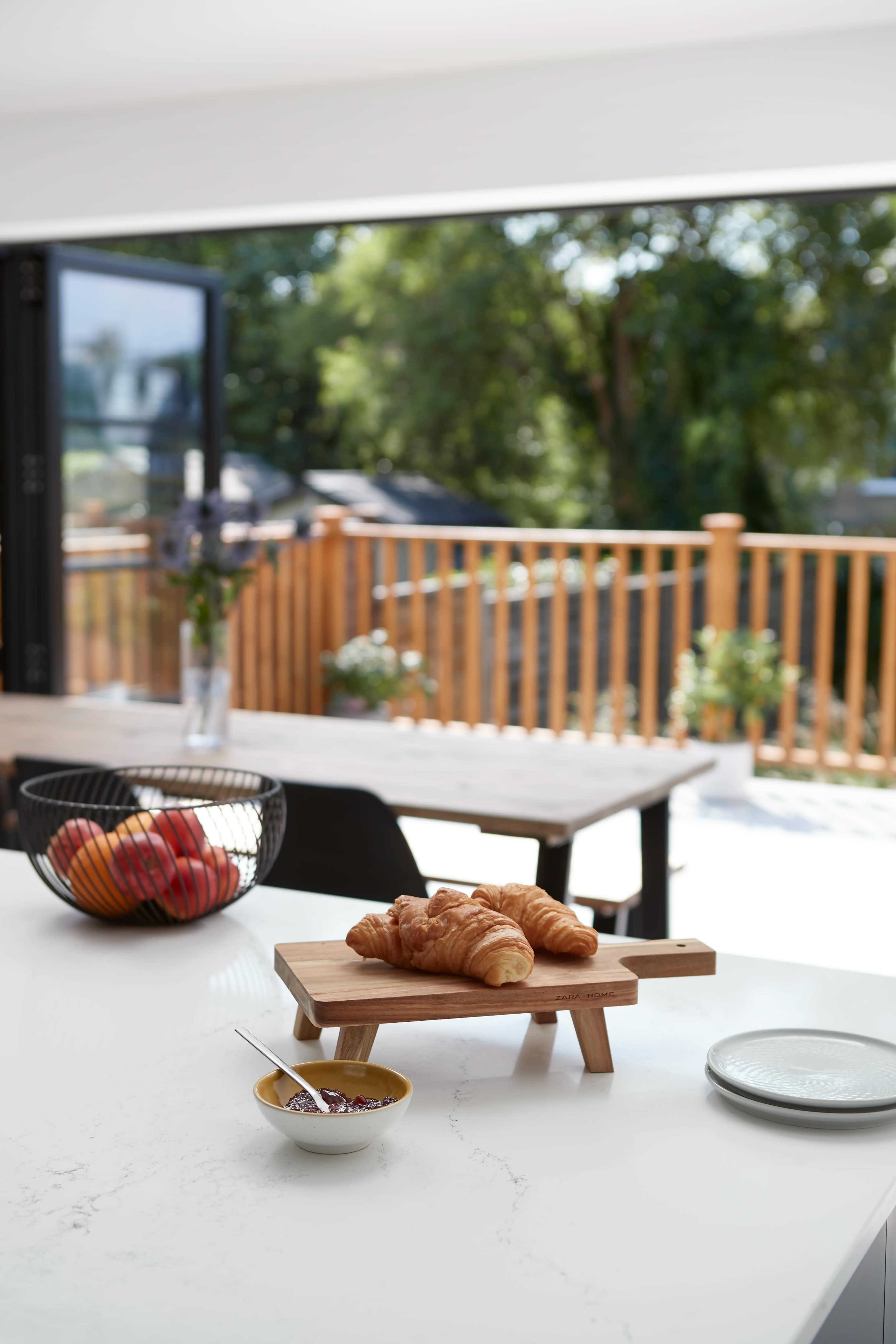 A wooden table holds a plate of croissants with a bowl of jam, while a bowl of apples sits nearby and a view of an outdoor patio is visible through large windows.