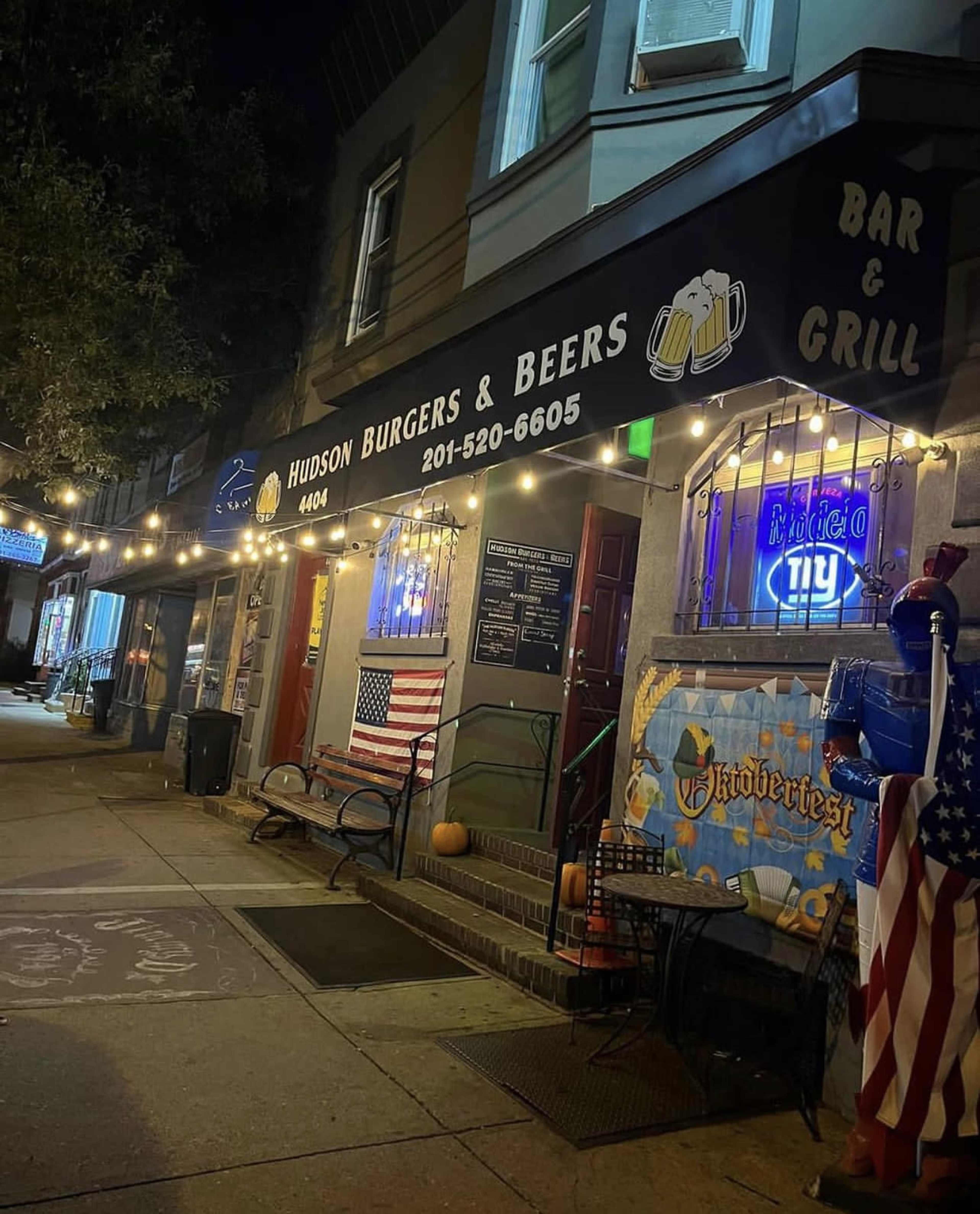 The image shows a bar and grill named "Hudson Burgers & Beers," featuring outdoor seating and a decorative storefront with string lights.