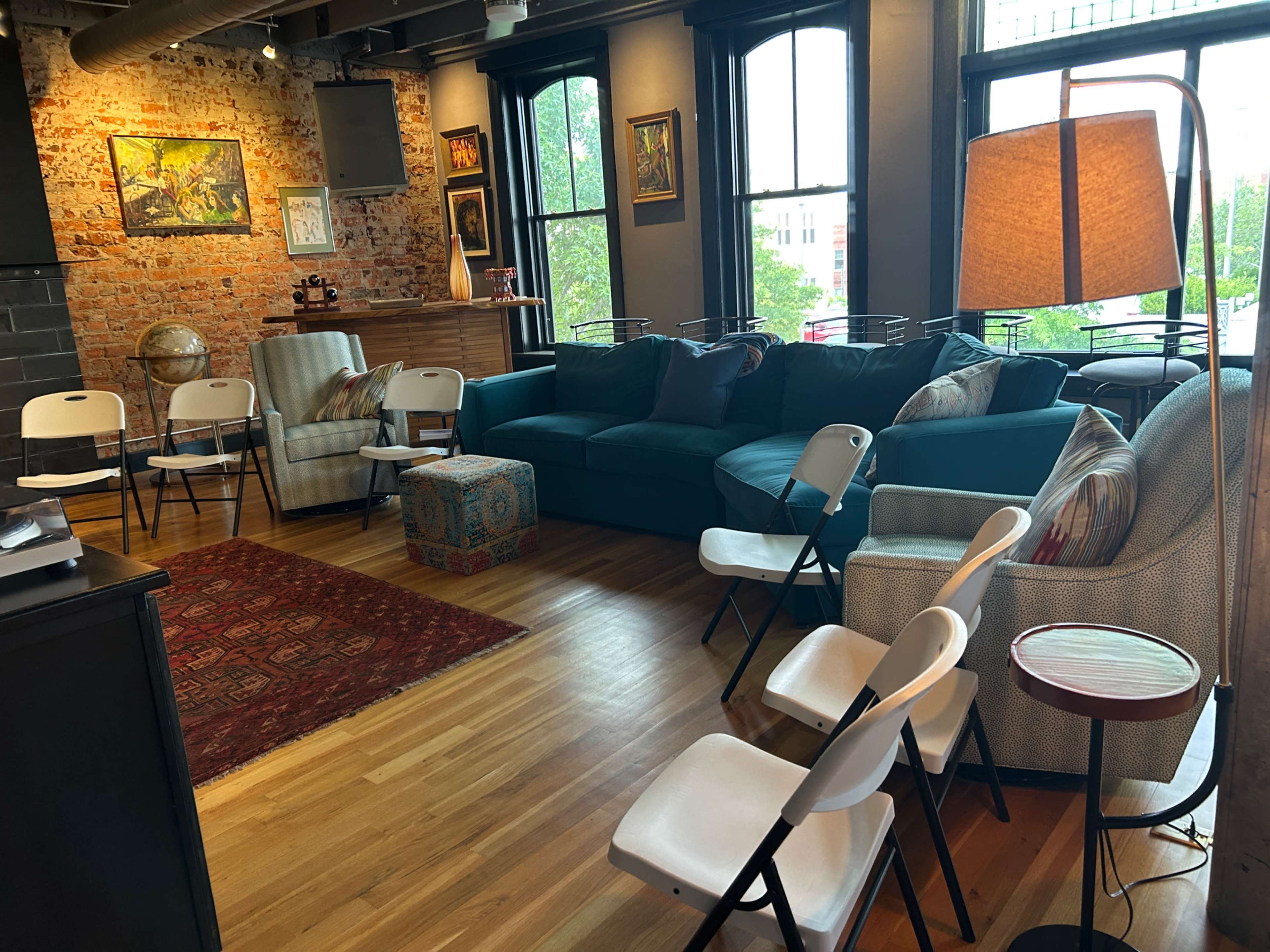 The image shows a cozy room with exposed brick walls, a blue sofa, several folding chairs arranged in a semi-circle, and a lamp beside a small side table.