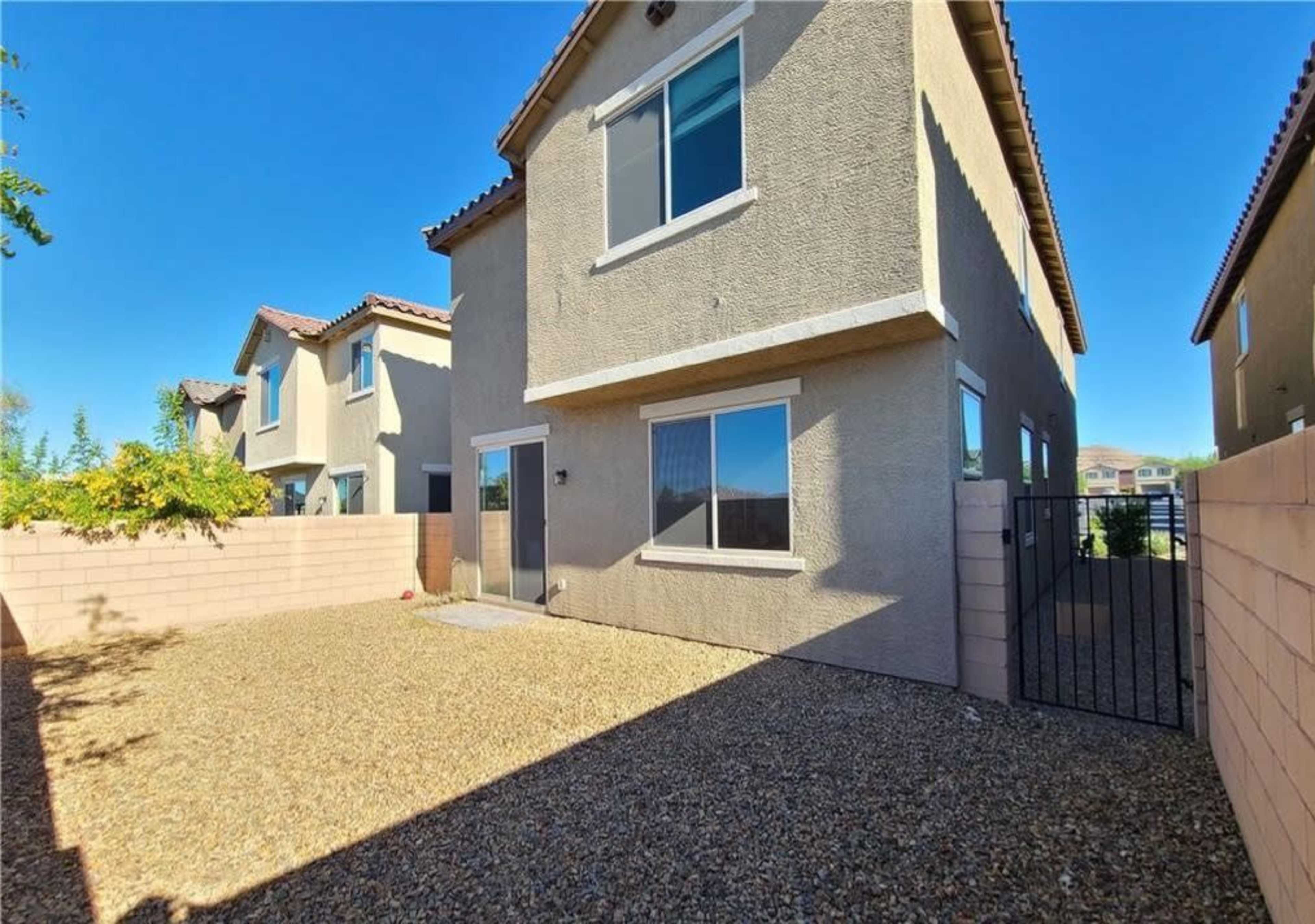 A two-story house with beige exterior walls and several windows is surrounded by a gravel courtyard and enclosed by a pink fence.