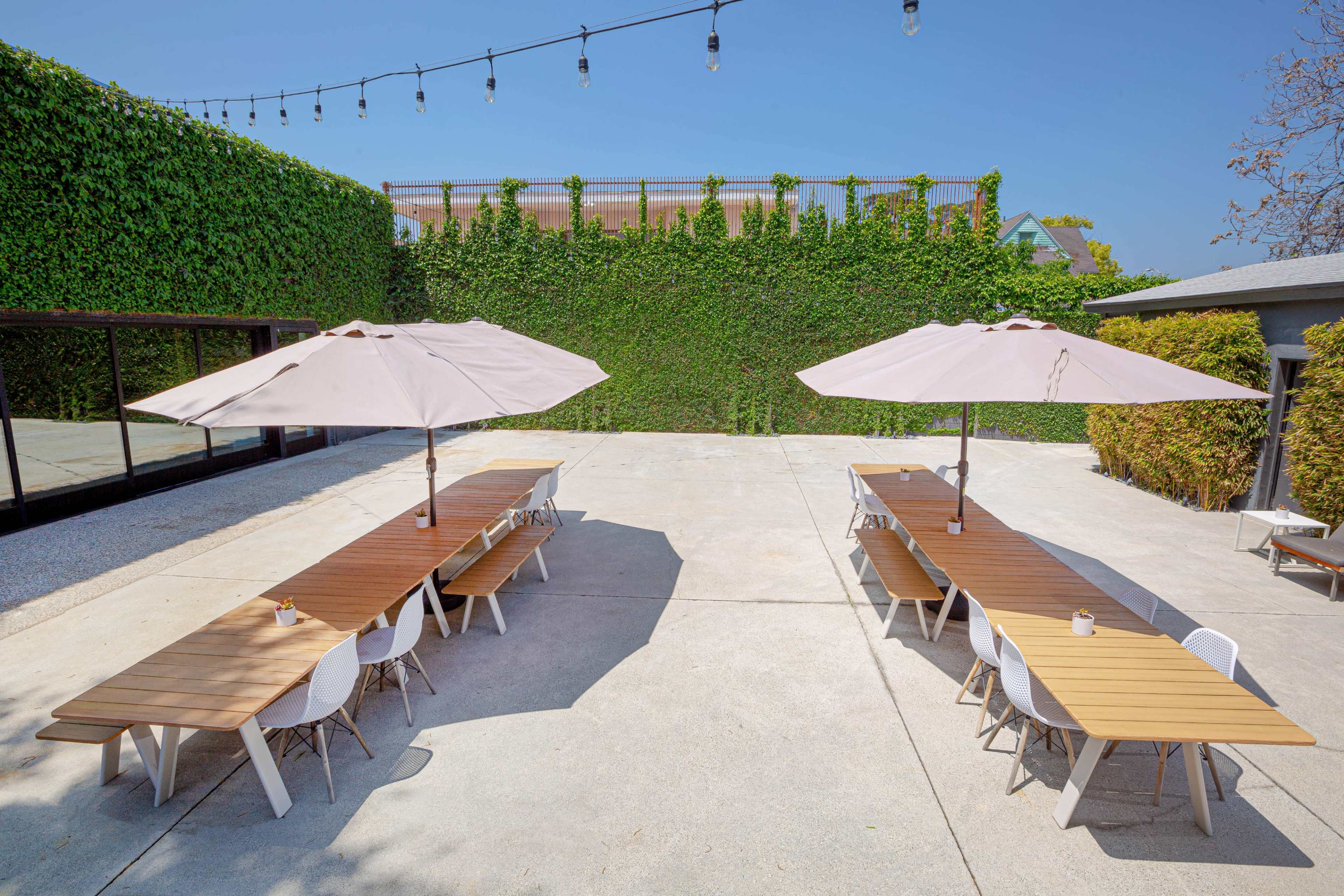 An outdoor dining area with two long wooden tables surrounded by white chairs, shade umbrellas, and a green vine-covered wall in the background.