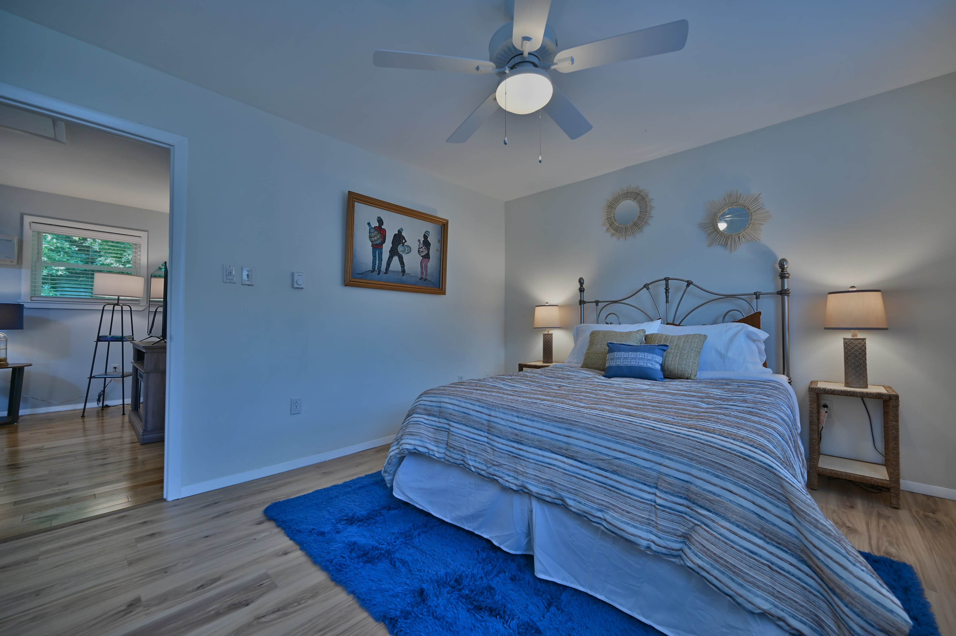 A bright, minimalist bedroom with a bed covered in striped bedding, a blue area rug, and framed artwork on the wall.