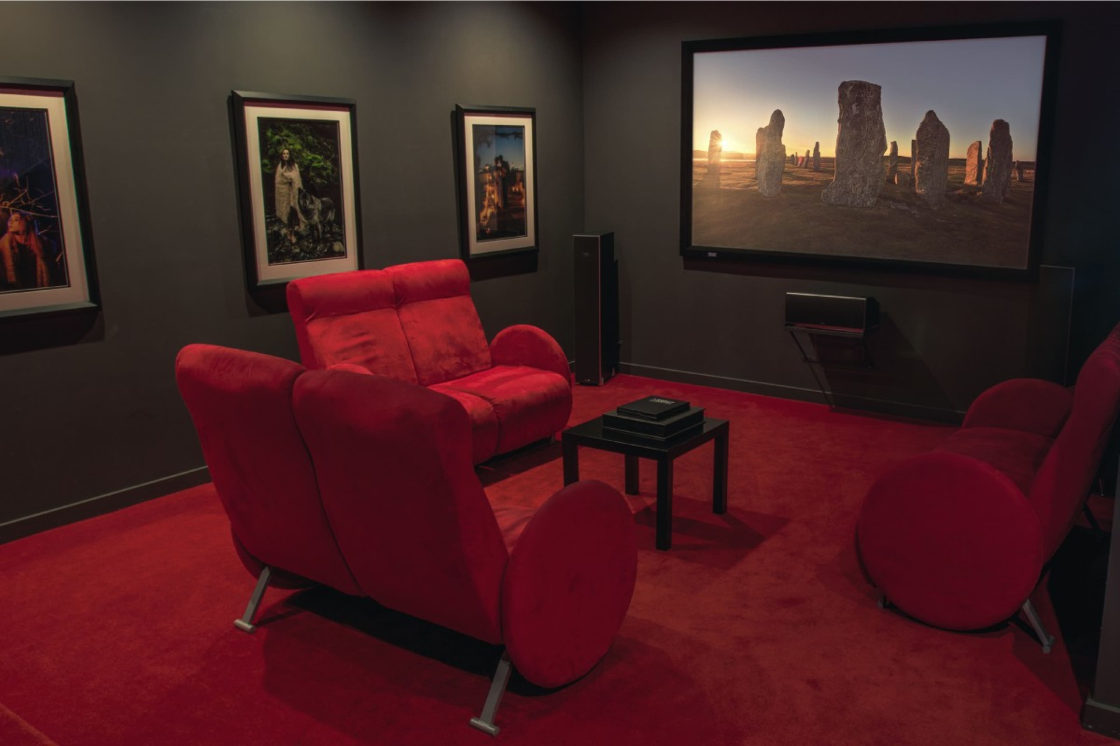 A home theater room with red seating, dark walls, and a large screen displaying stone formations at sunset, surrounded by framed artwork on the walls.