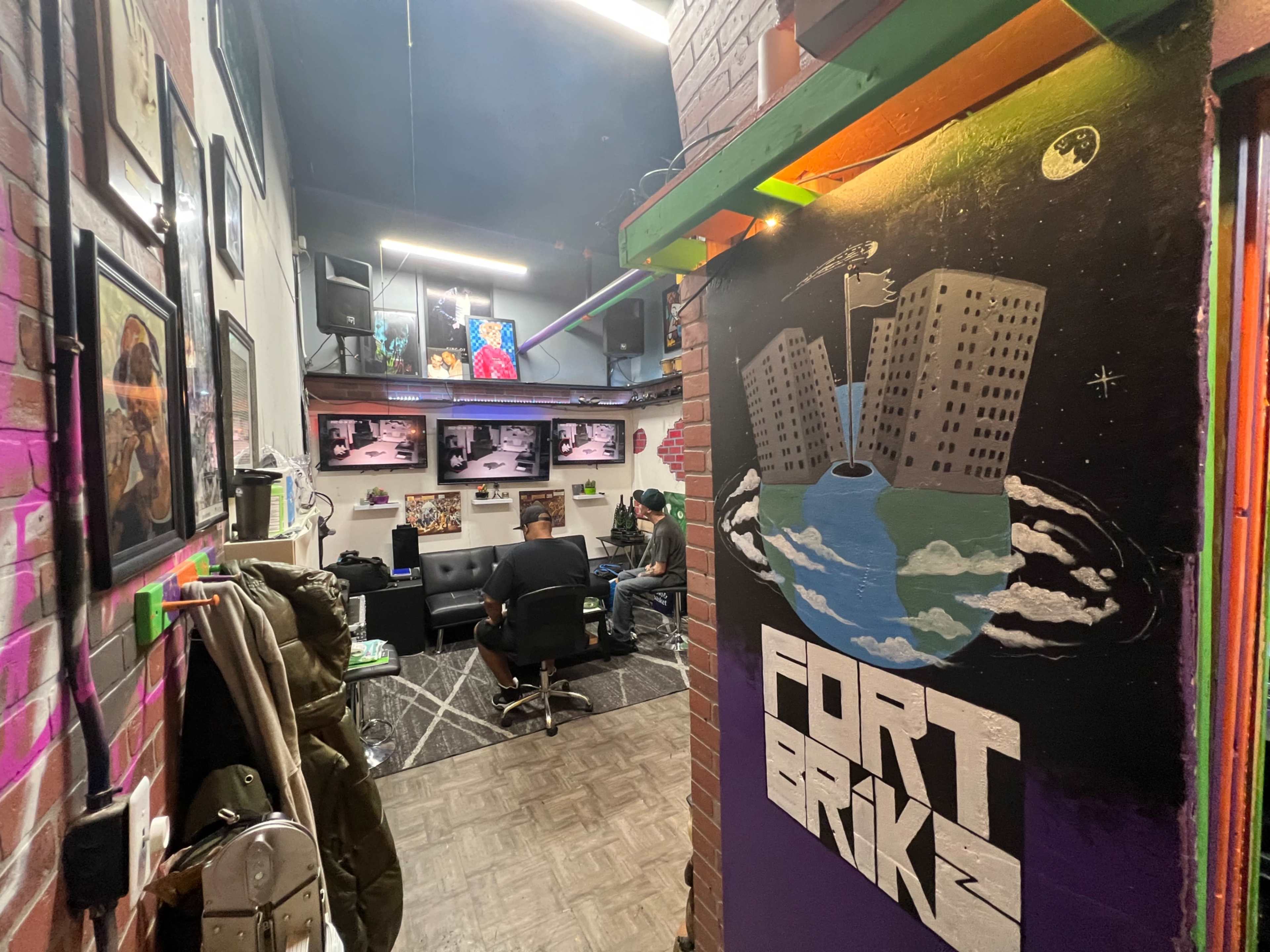 An interior space with a colorful mural on one wall, featuring the words "FORT BRIK," and several television monitors displaying images, while a person sits at a desk in the background.