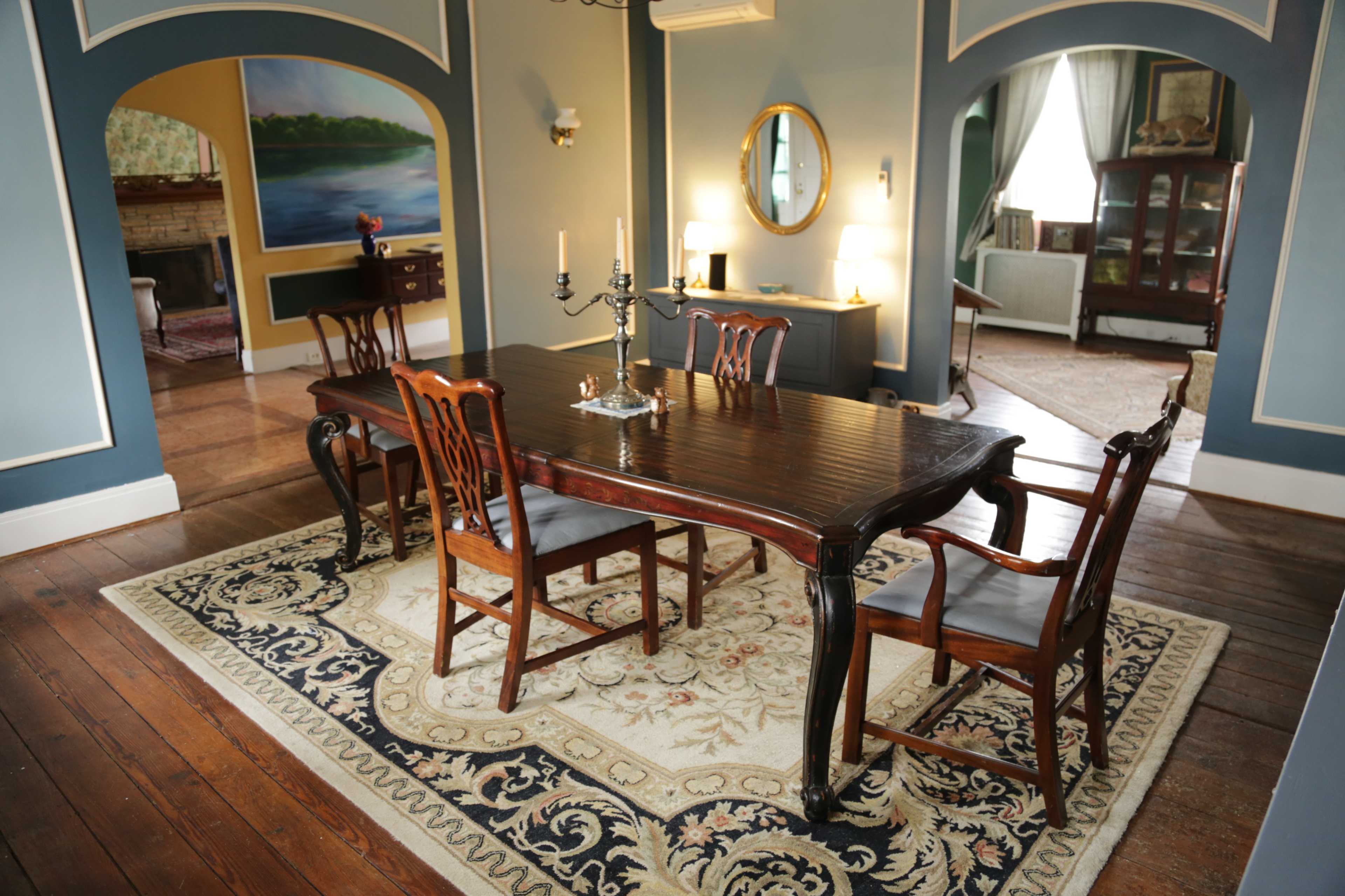 A dining room with a large wooden table surrounded by several chairs, set on a patterned rug, with light blue walls and a painting in the background.