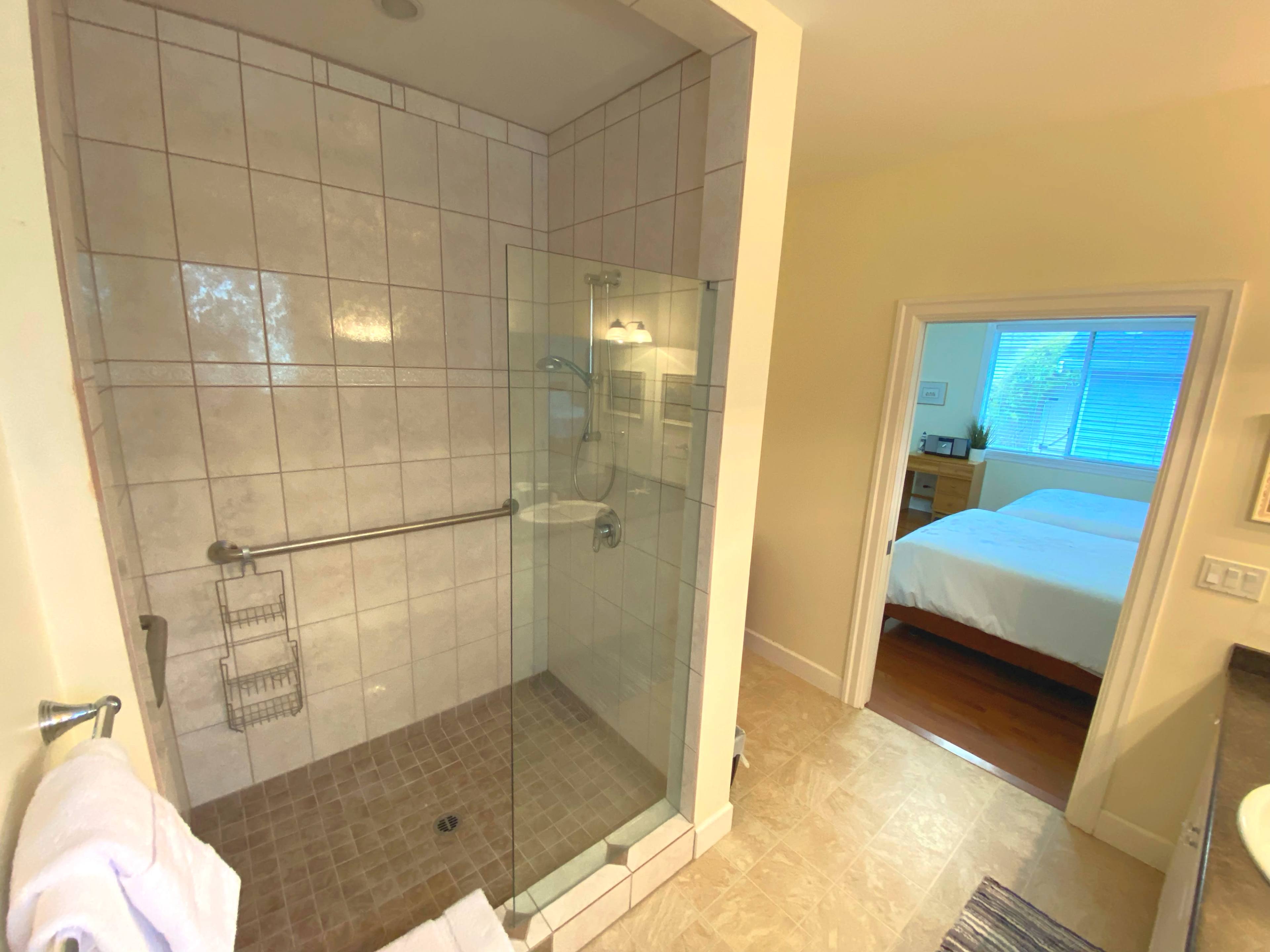 A corner shower with glass walls and a tiled floor adjacent to a doorway leading to a neatly arranged bedroom.