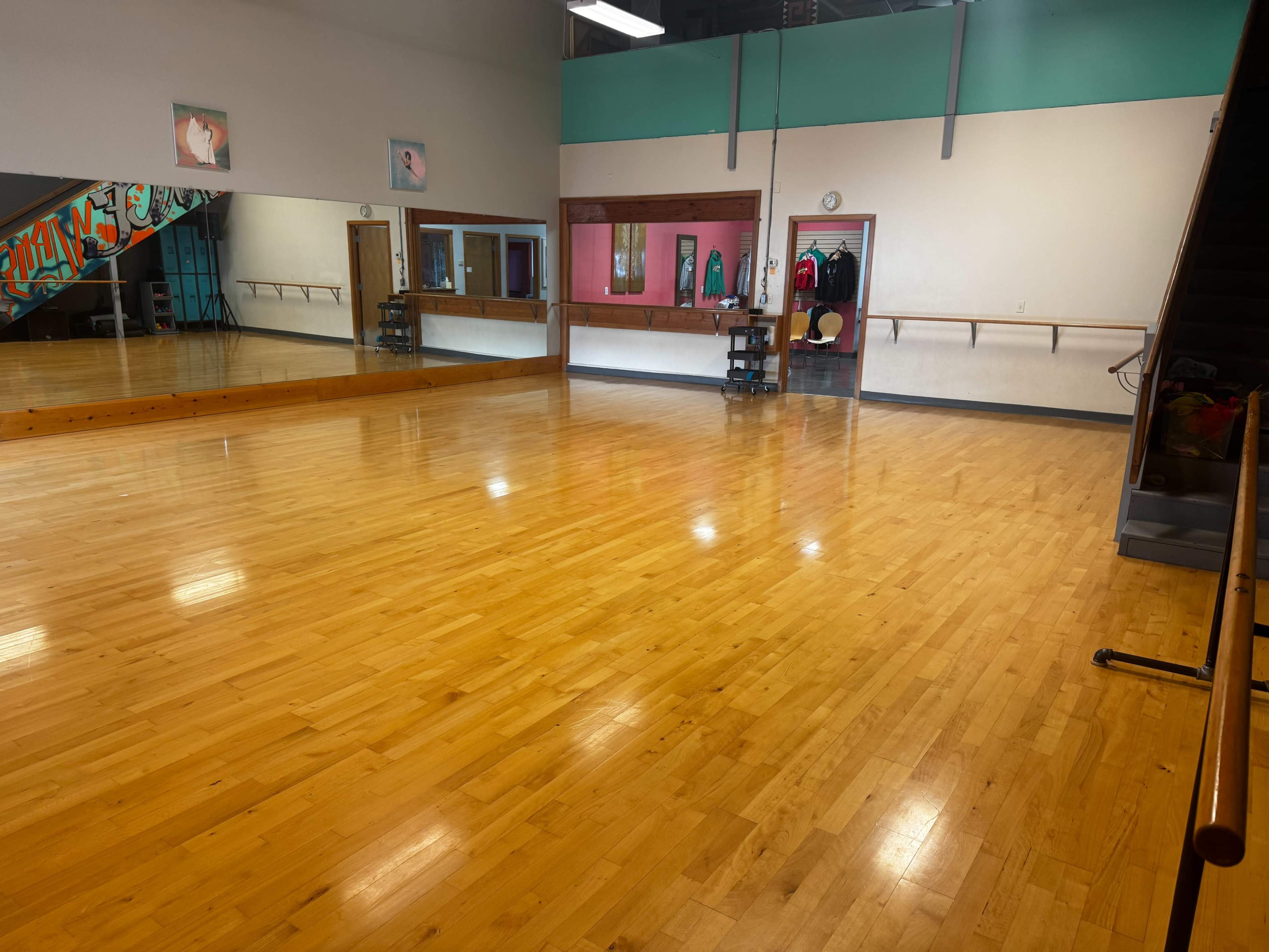 The image shows a spacious dance studio with polished wooden floors, large mirrors on one wall, and a door leading to another room at the back.