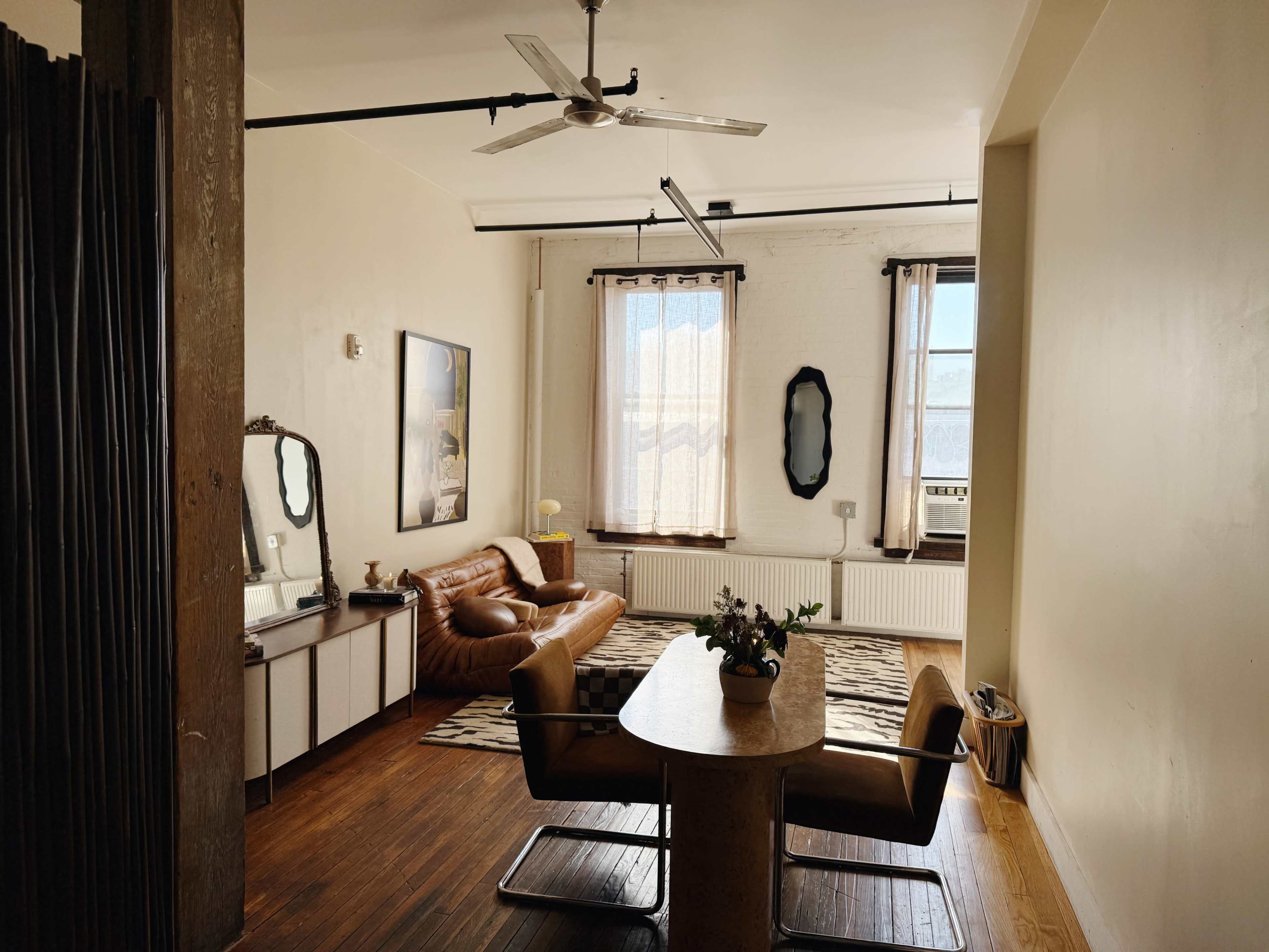 The image shows a spacious living area with a brown leather sofa, a round dining table with chairs, and large windows allowing natural light to enter.