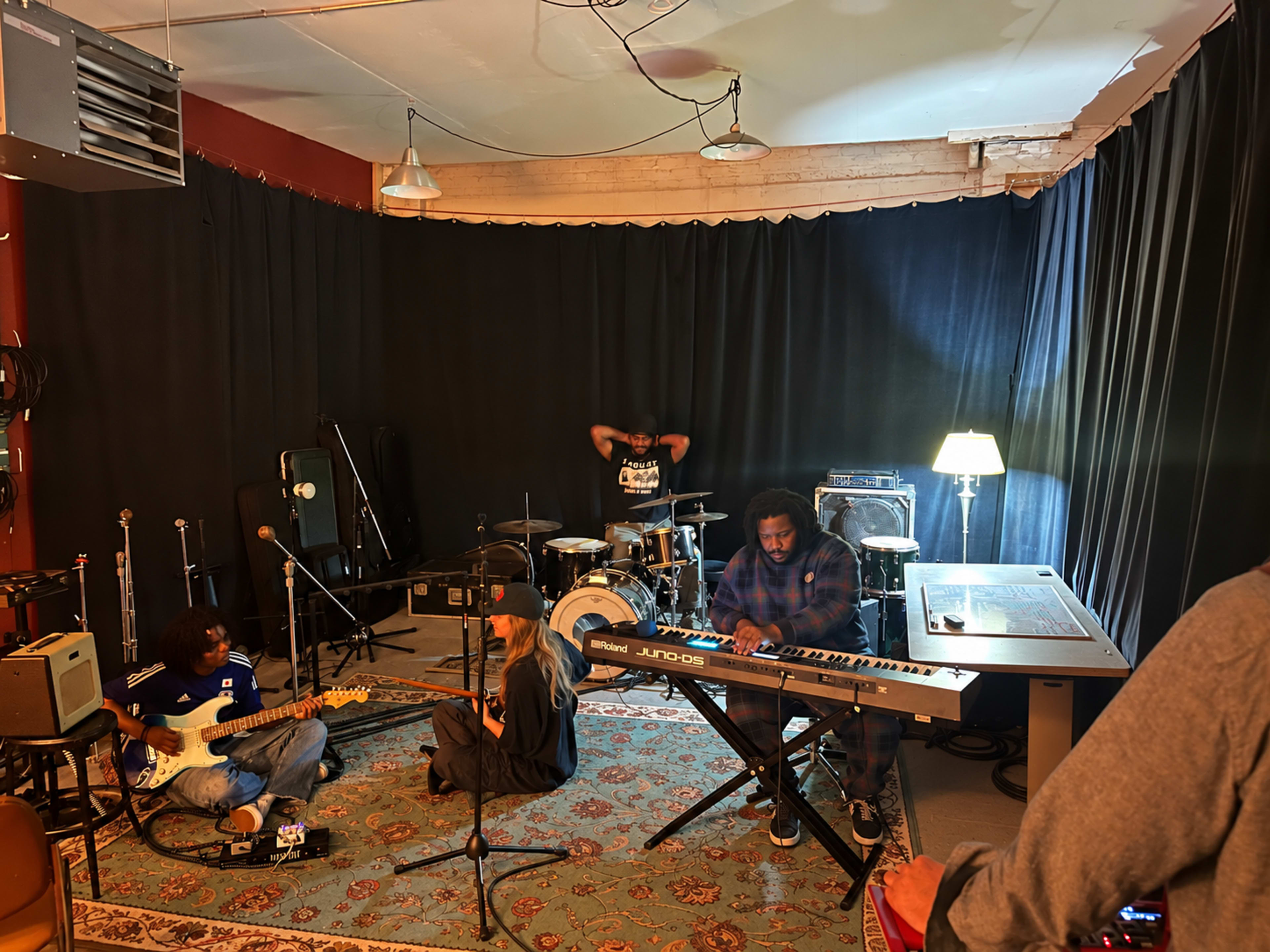 A group of musicians is practicing in a soundproof room with instruments including a guitar, keyboard, and drums, while one member is seated on the floor and another is at the piano.
