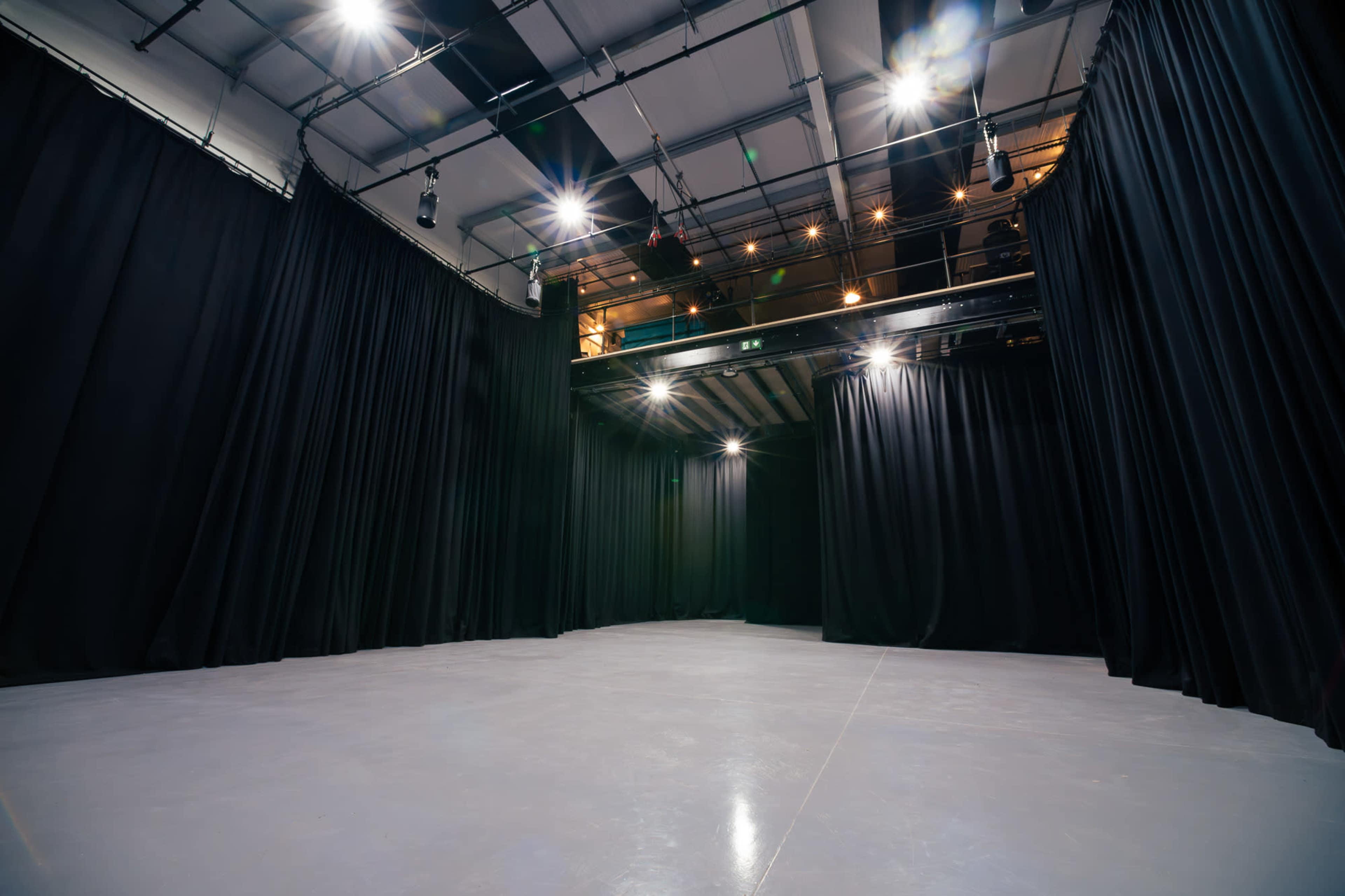 An empty theater stage with black curtains and overhead lighting.