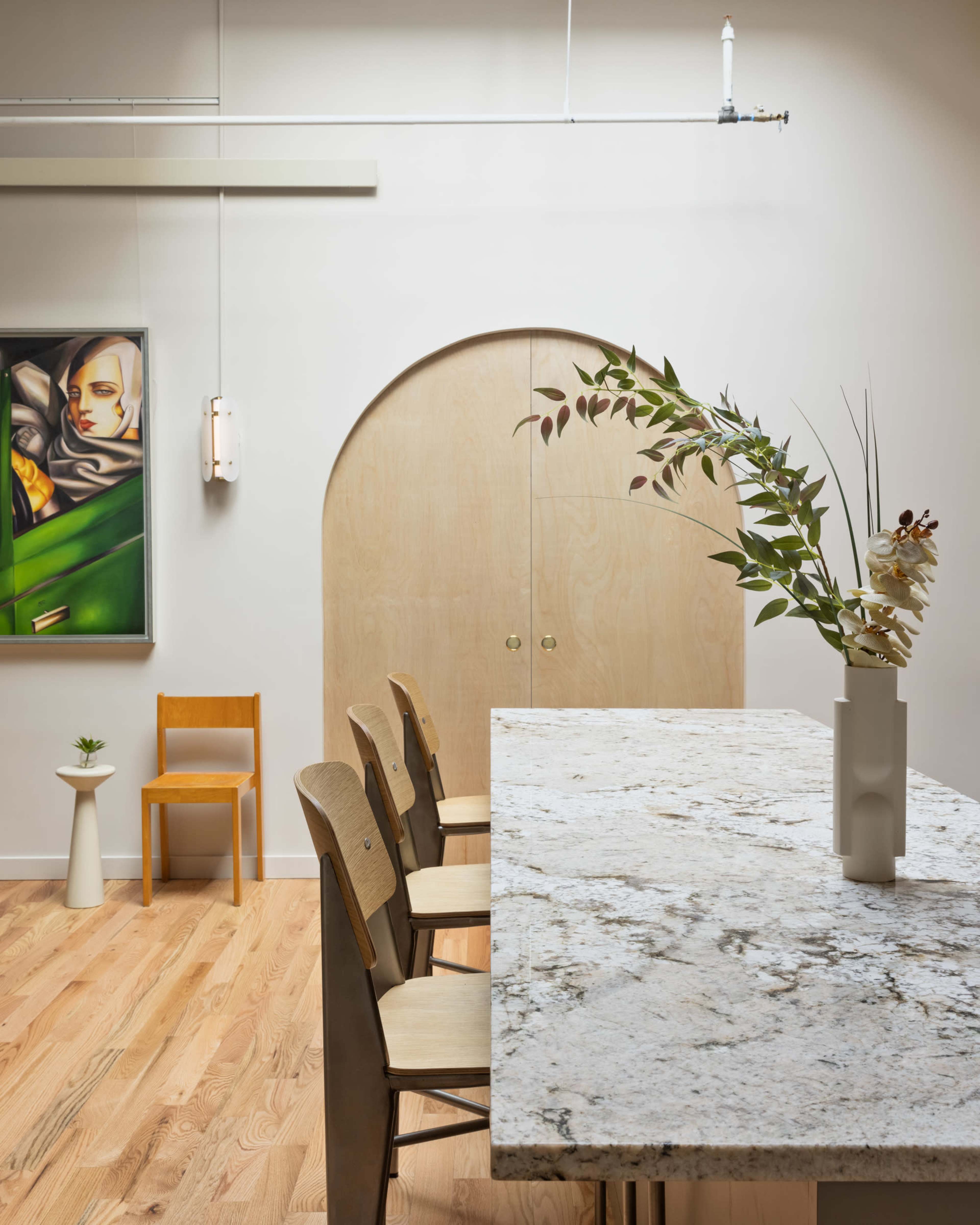 A modern interior features a large granite table with wooden stools, a minimalist chair, and a decorative vase with flowers, all set against a backdrop of artistic decor and wood finishes.