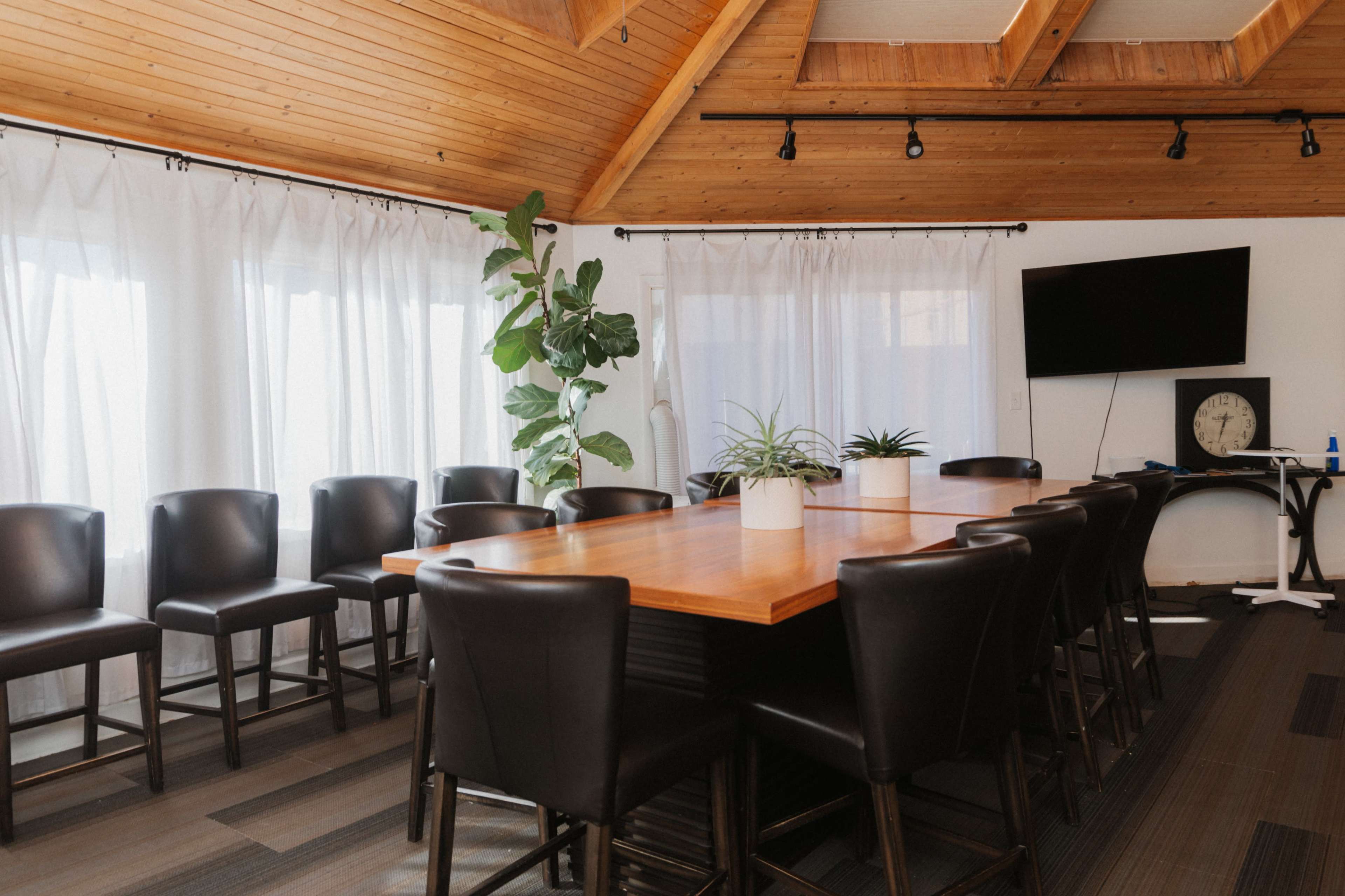 A large conference room features a wooden table surrounded by black chairs, with plants and large windows covered by sheer curtains.