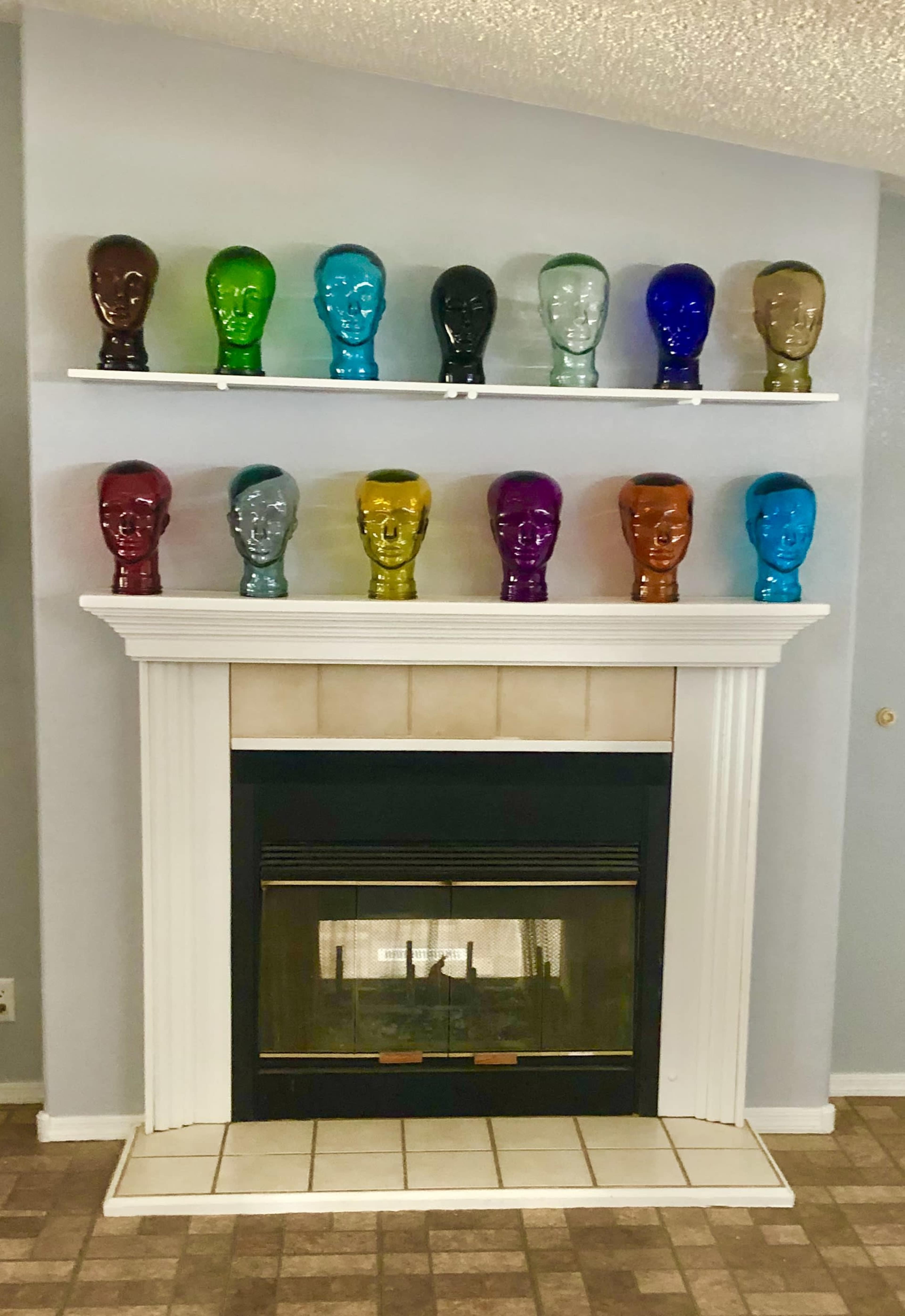 A row of colorful glass head sculptures is displayed on a shelf above a white fireplace in a room with gray walls.