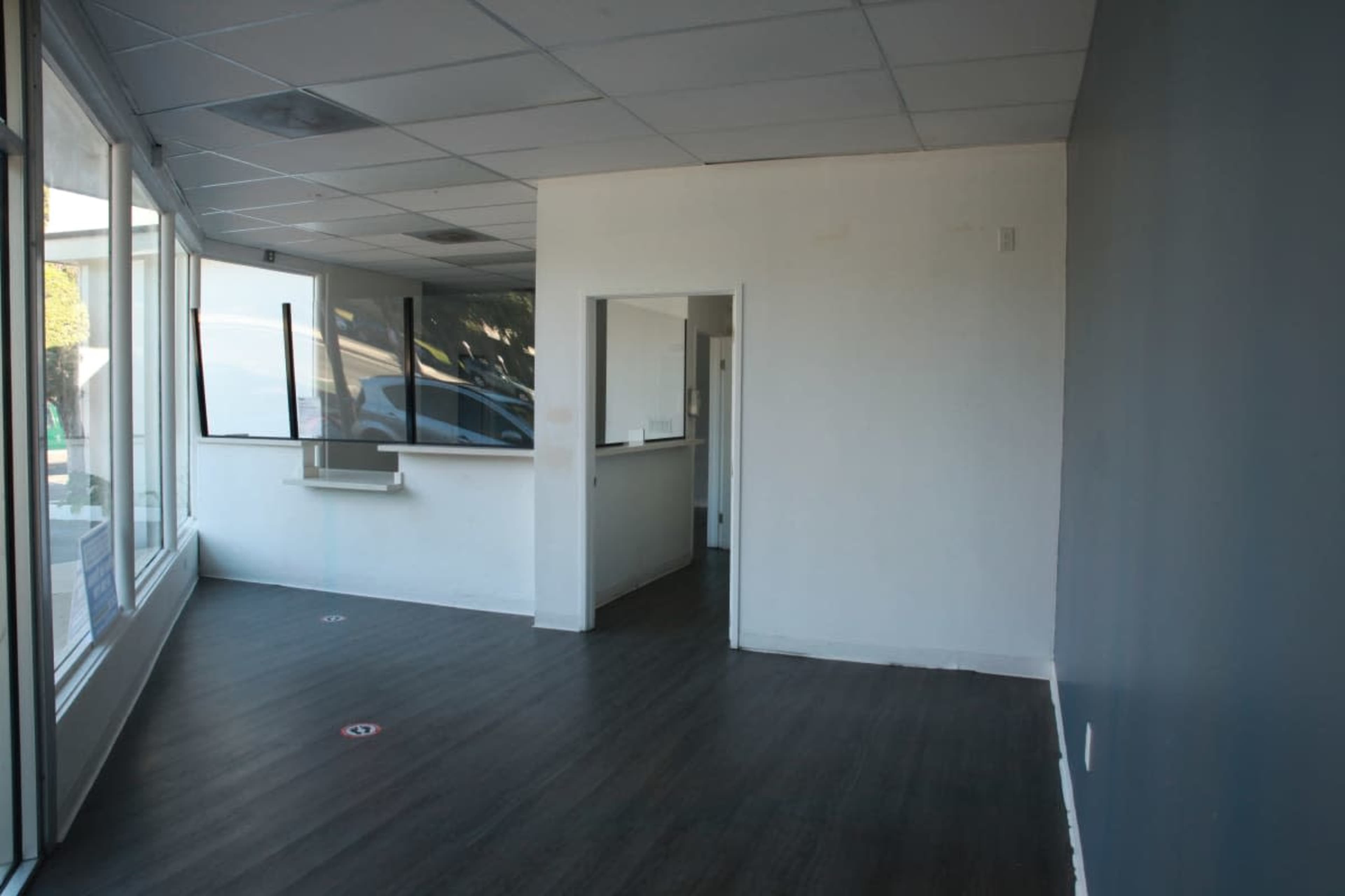 The image shows an empty interior space with a large window, dark wooden flooring, and a partially visible room in the background.