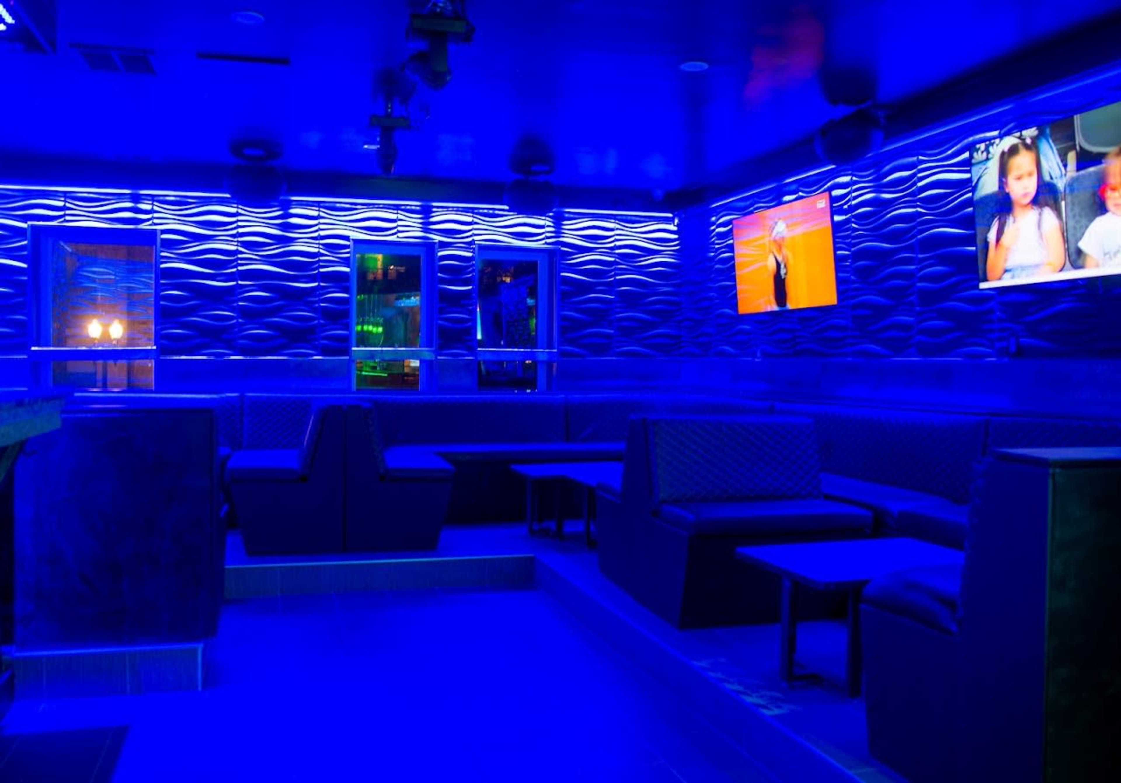 The image shows a dimly lit lounge area with blue lighting, featuring benches and tables, along with screens displaying videos on the walls.