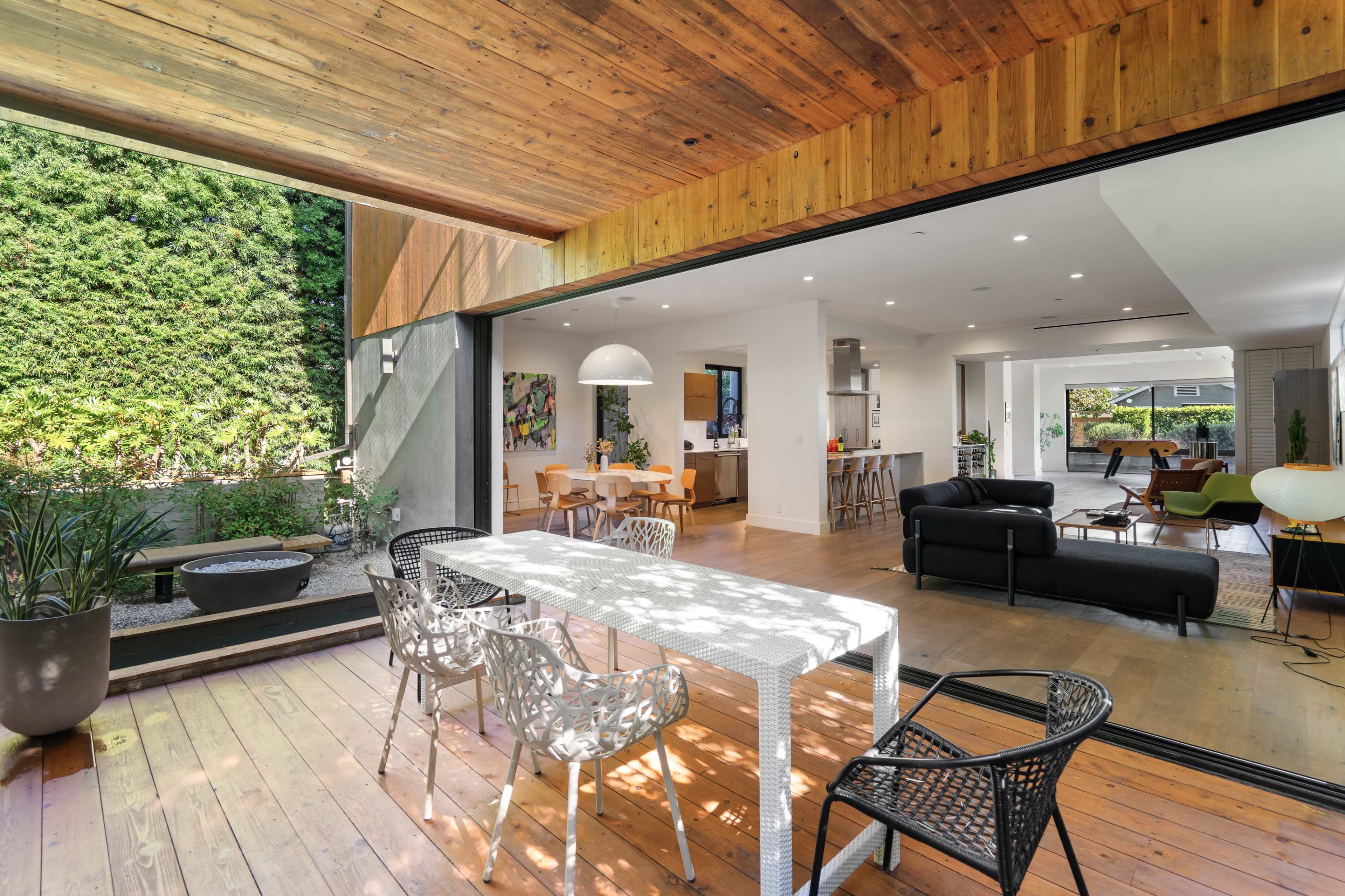 A modern open-concept living space features a seamless transition from an indoor dining area to an outdoor patio, surrounded by greenery.