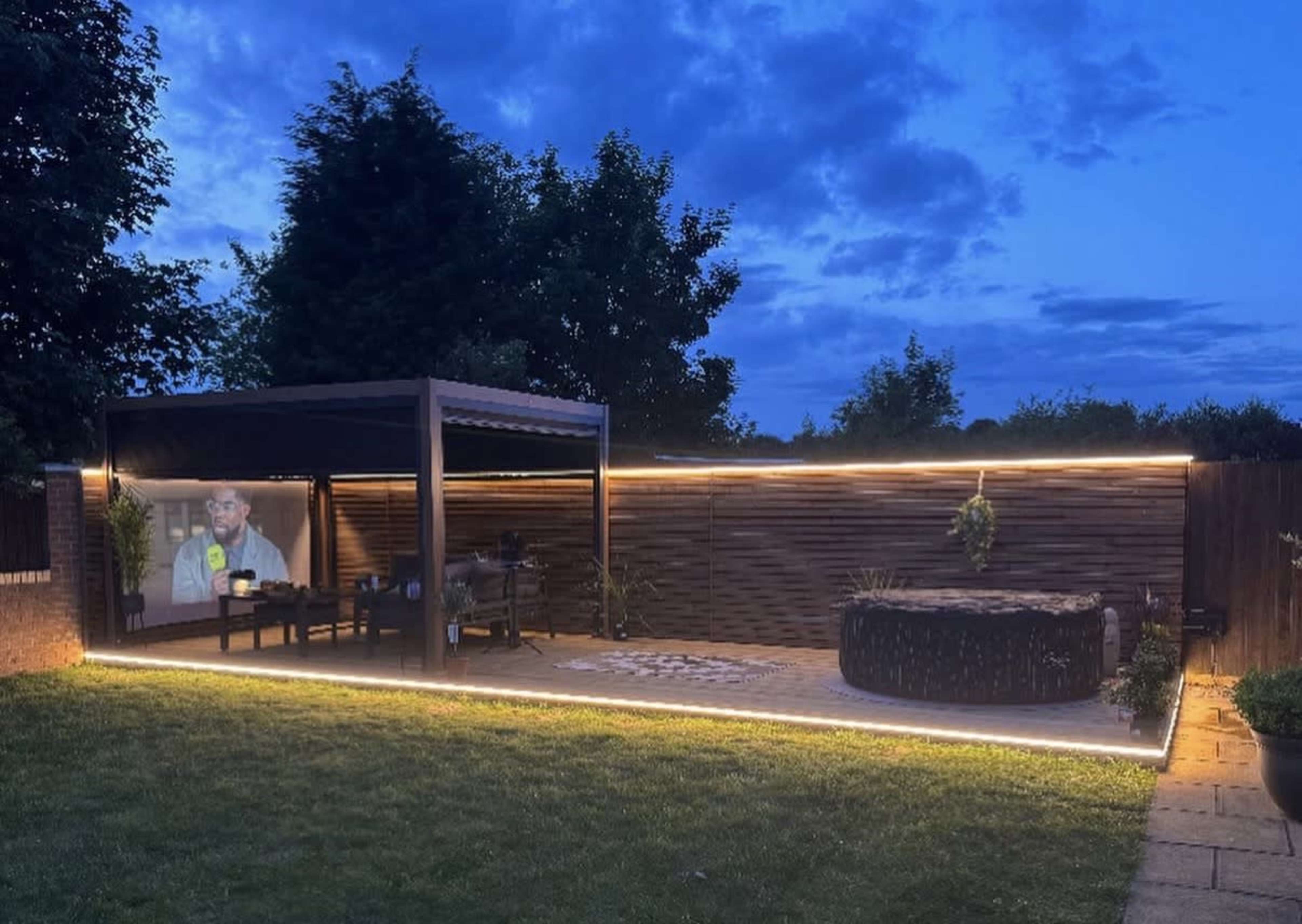 The image shows a backyard with a modern pergola, a seating area, a hot tub, and ambient lighting along the wooden fence, set against a twilight sky.