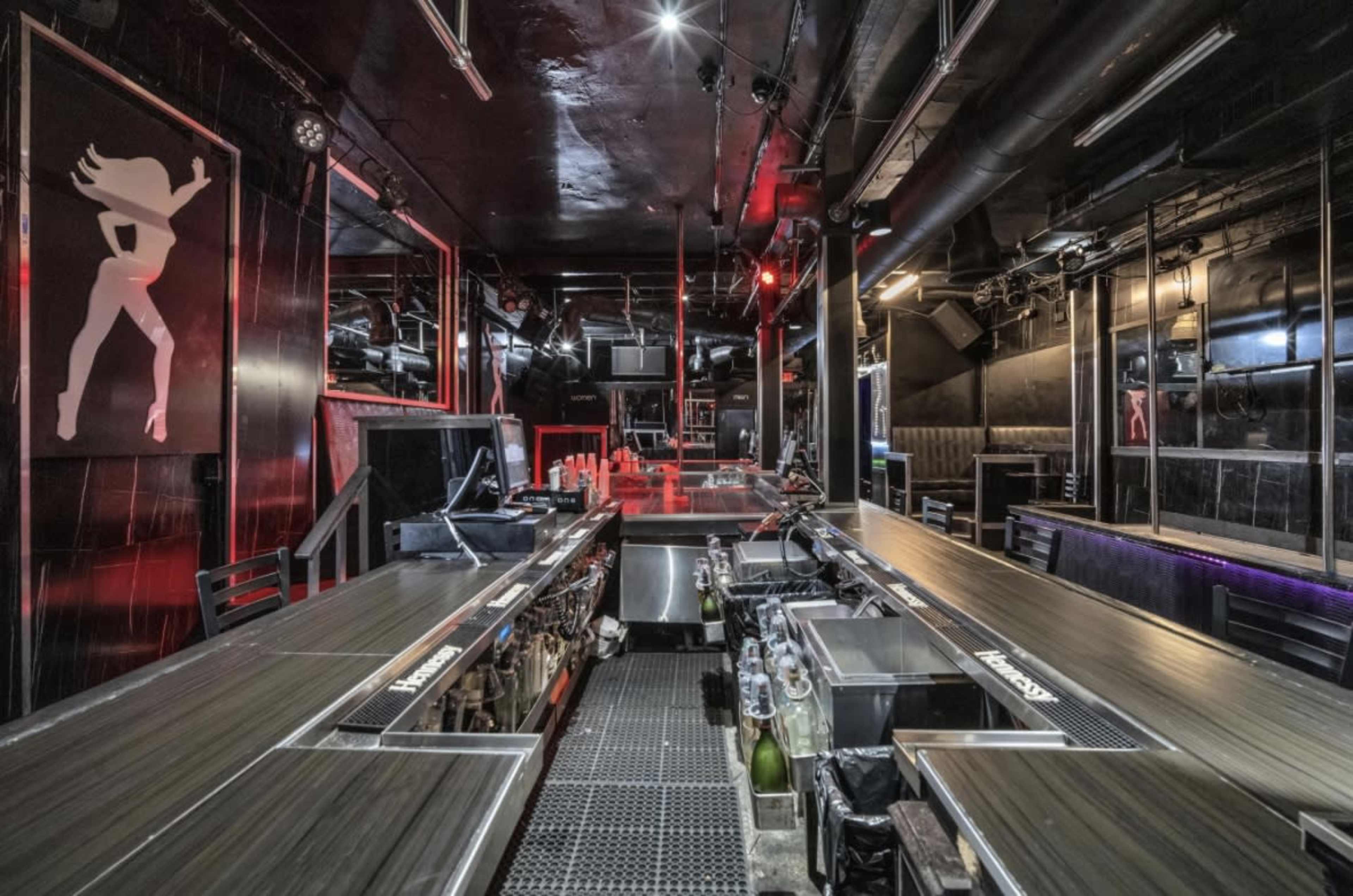 The image shows a dimly lit bar interior with a long countertop, metal shelving, and a decorative wall featuring a silhouette of a dancer.