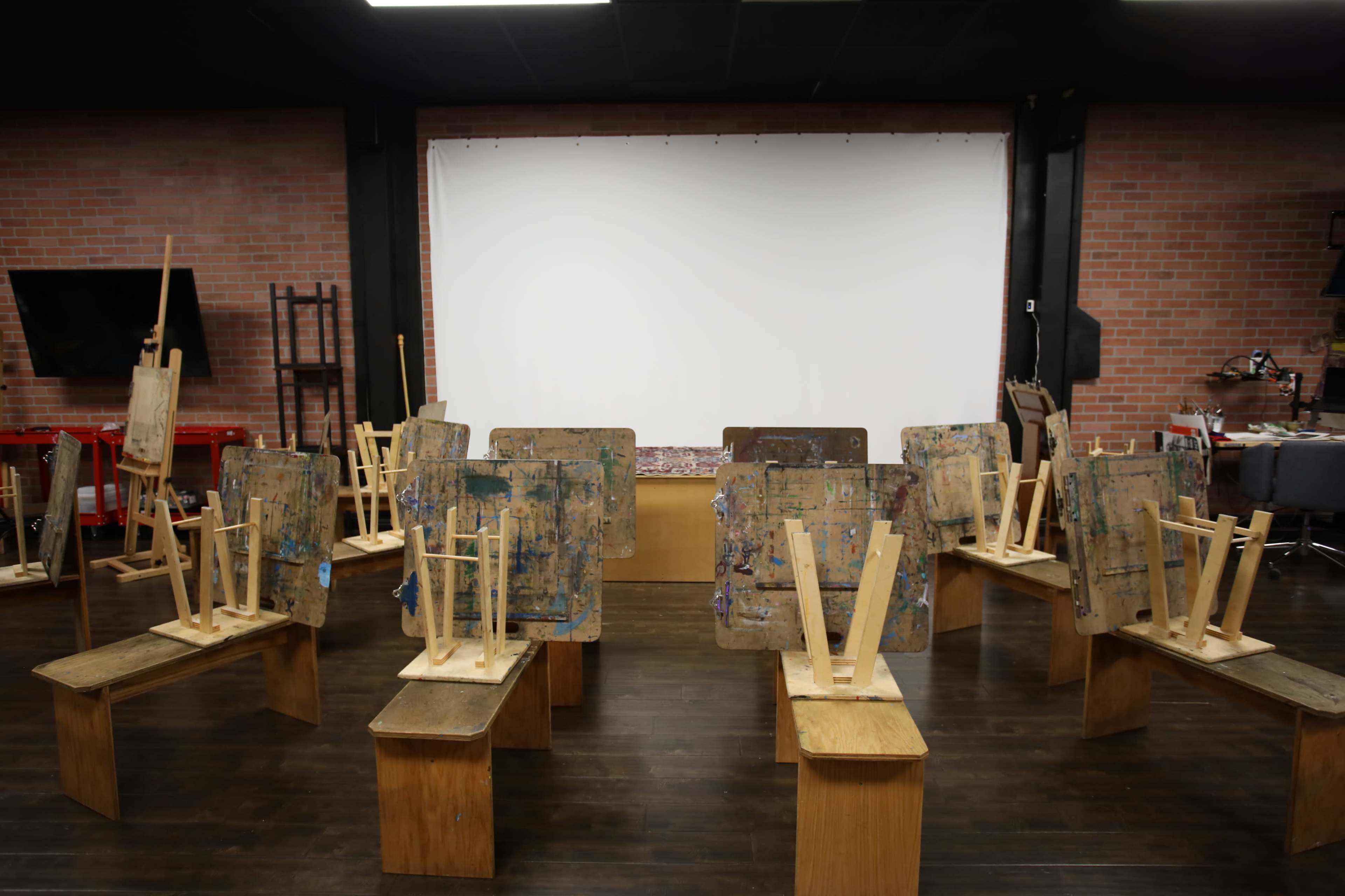 A room with several wooden easels turned upside down on wooden benches, facing a blank white screen at the front.