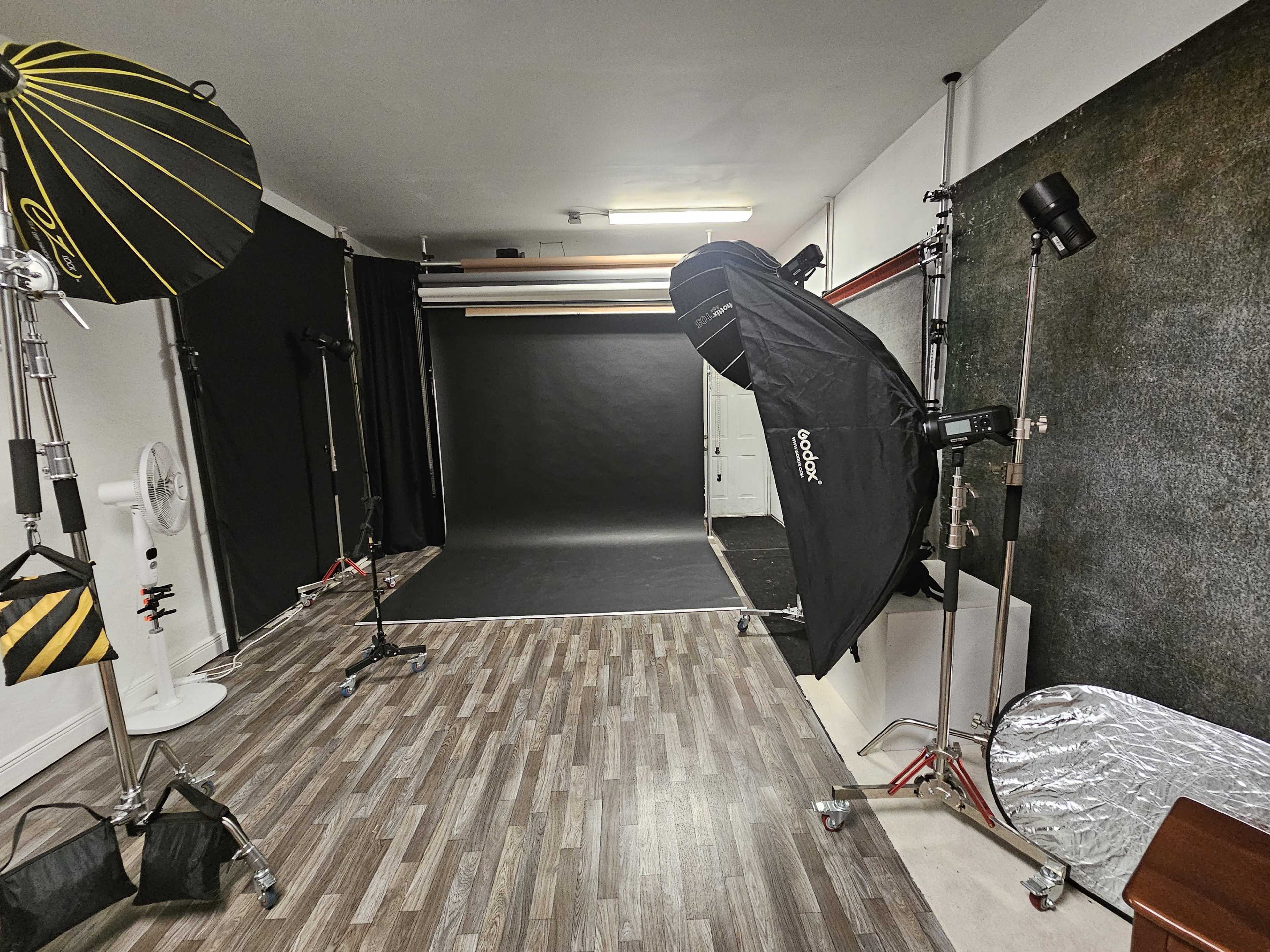 The image shows a photography studio equipped with lighting setups, a dark backdrop, and wooden flooring.