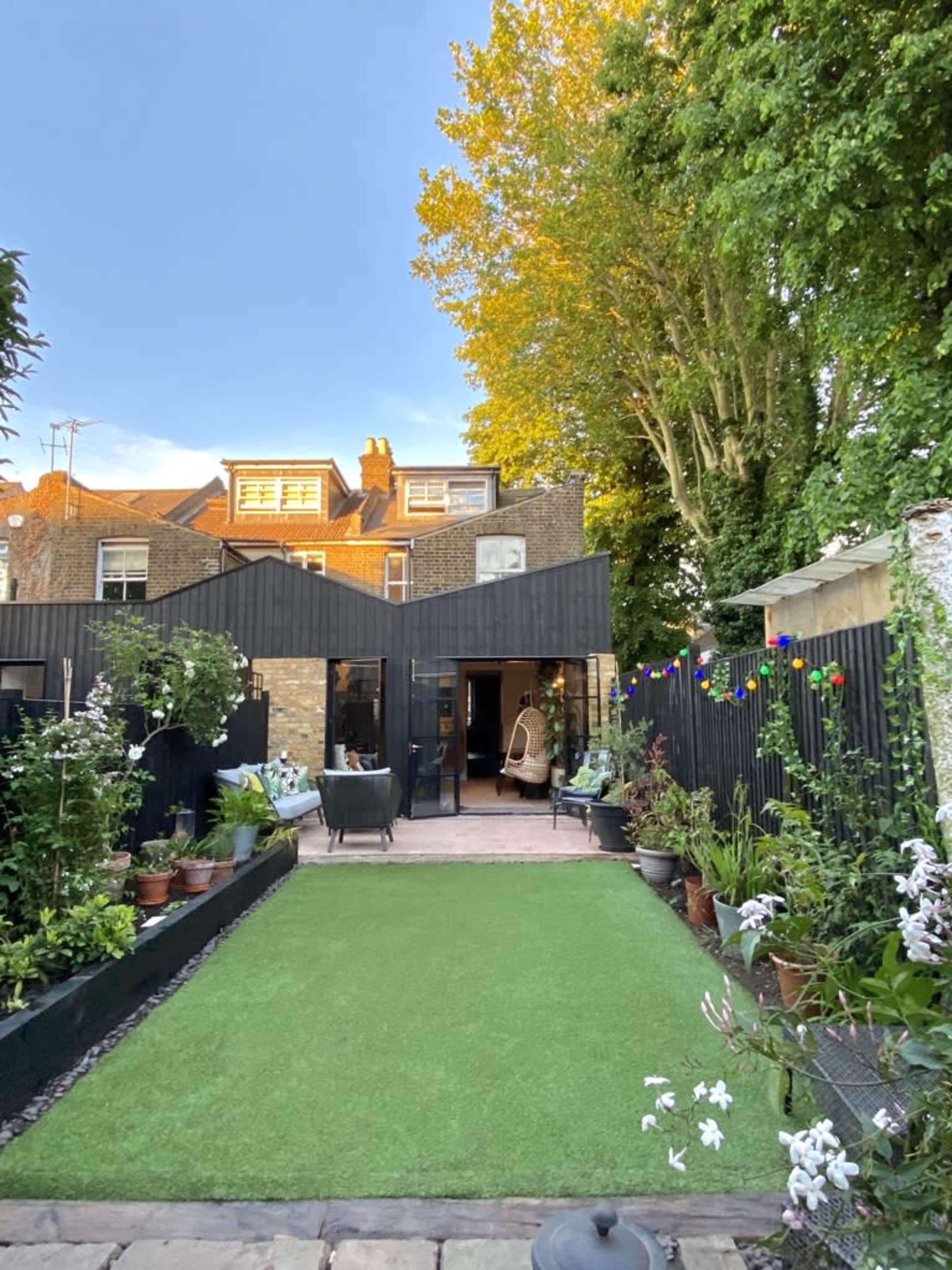 A well-maintained garden features green artificial grass, potted plants, and a modern black shed with large windows, set against a backdrop of a sunny sky and trees.