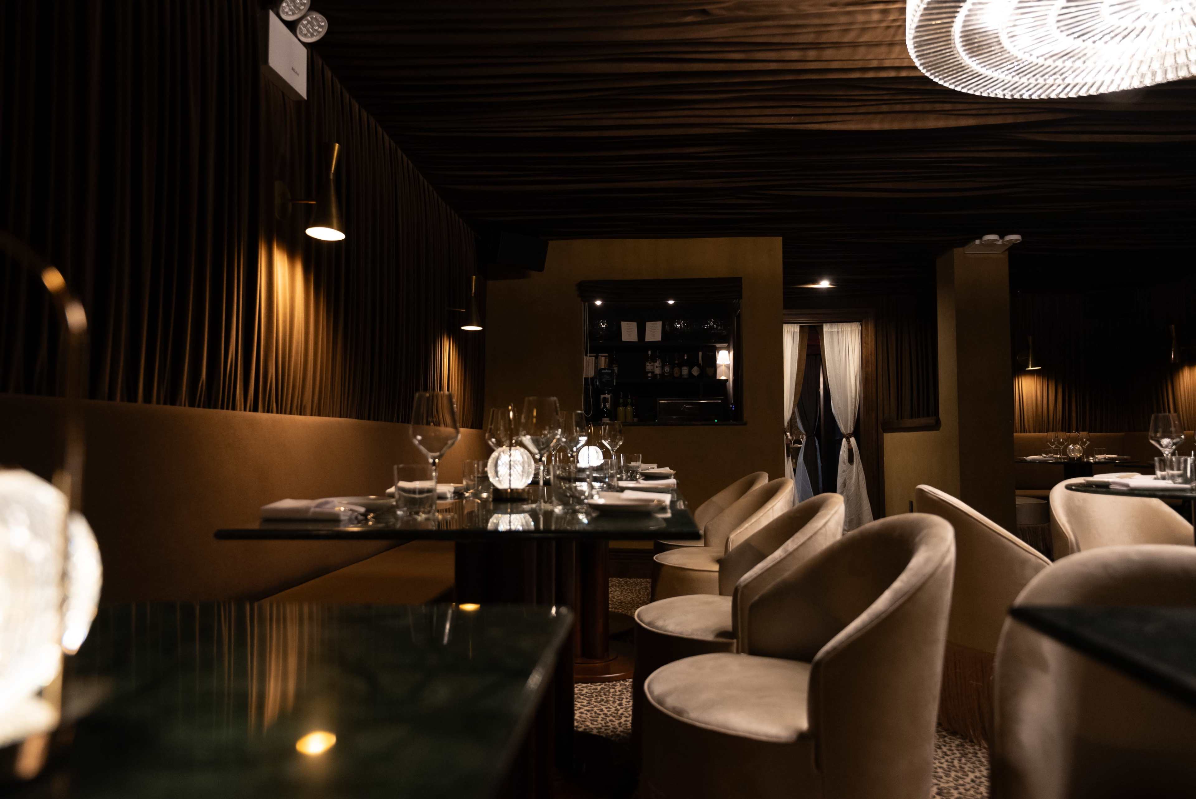 A dimly lit, elegant restaurant interior featuring plush seating, table settings with glassware, and rich, textured fabrics on the walls.