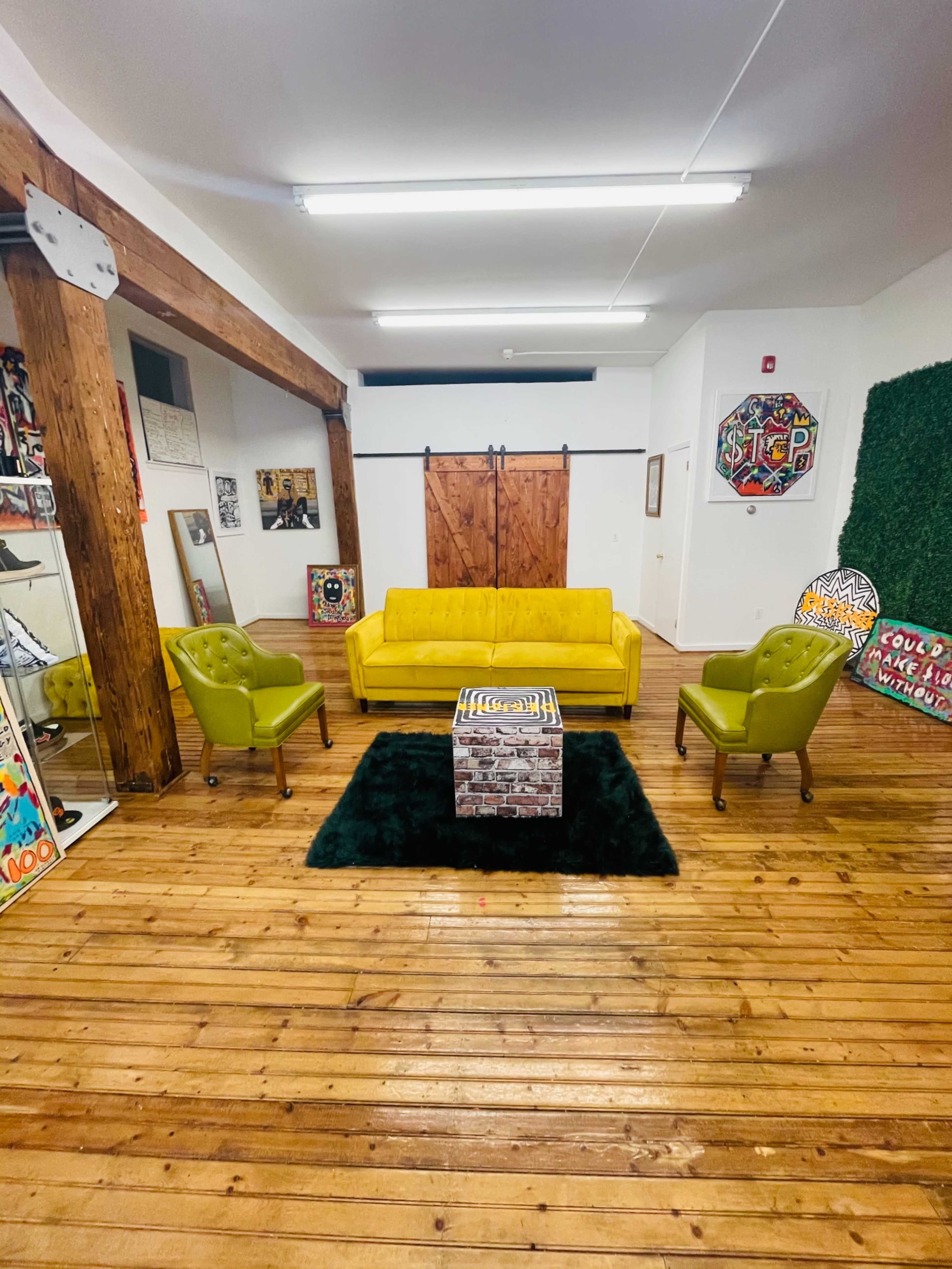 A bright room features a yellow sofa, two green armchairs, and a square coffee table on a dark rug, with wooden floors and exposed beams.
