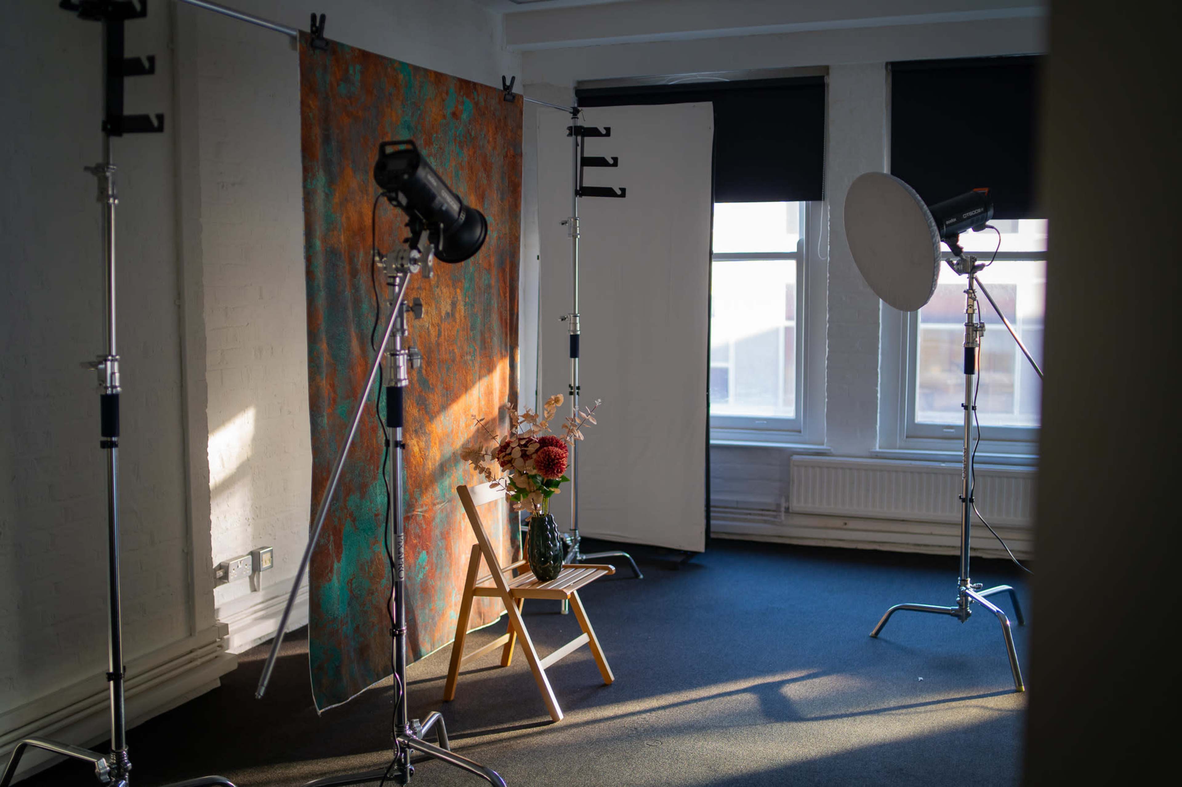 A photography studio with a backdrop, lighting equipment, and a vase of flowers on a chair.
